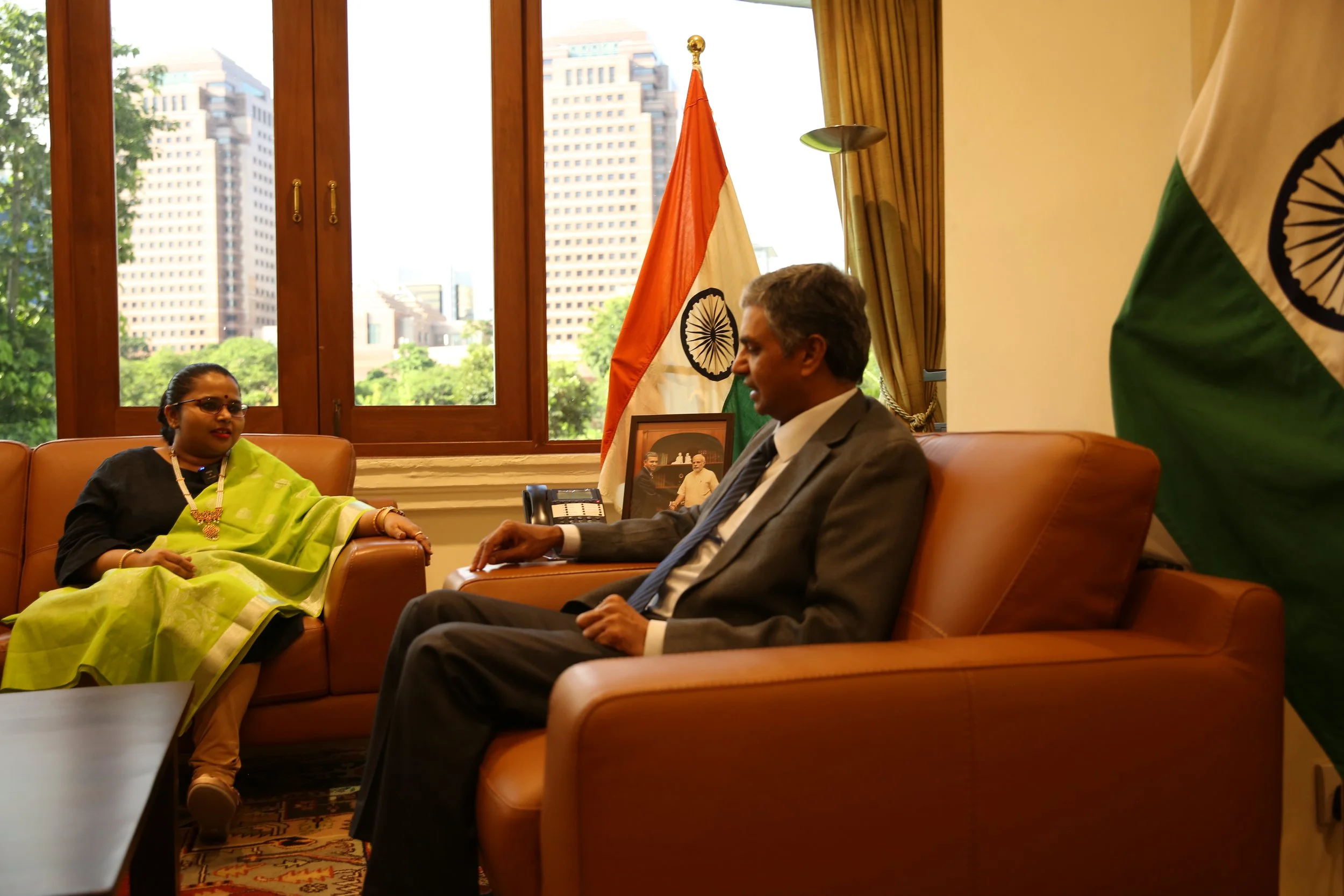 Two people sitting and talking in an office with Indian flags, a framed photograph, and a telephone on a desk. Large windows show city buildings and trees outside.