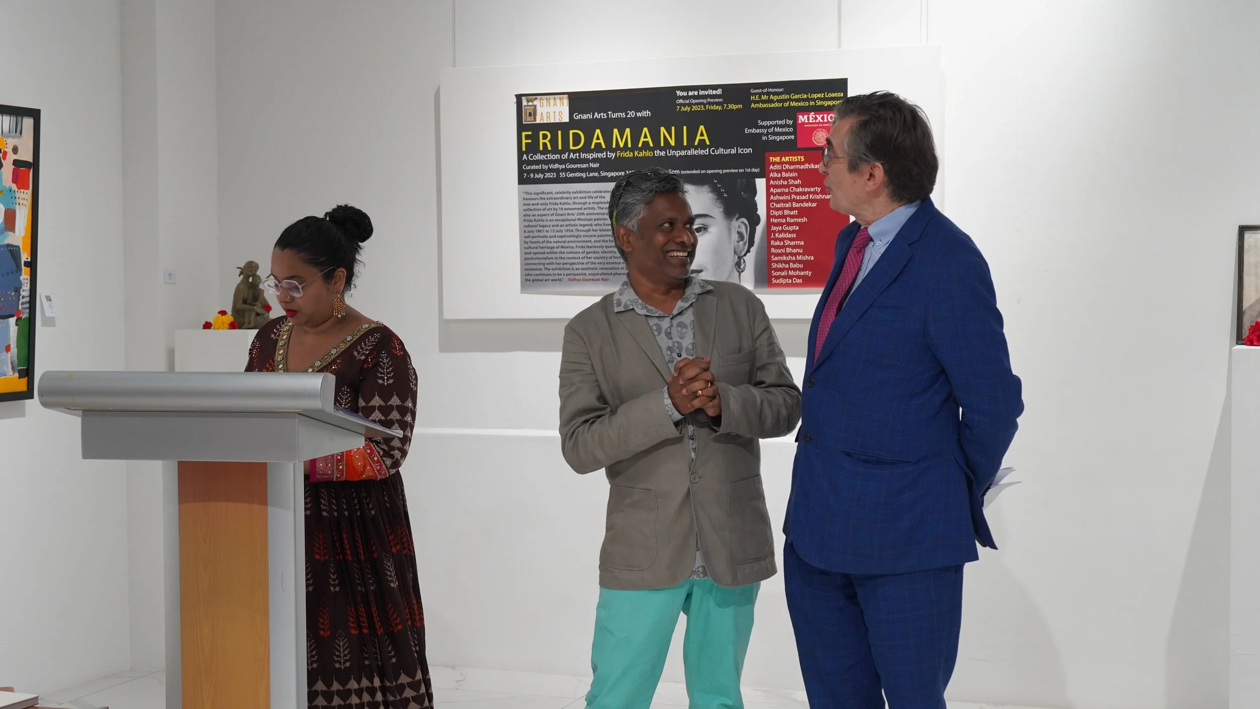 Three people in art gallery; woman at left wearing traditional dress, man in center with hands clasped, man in right wearing blue suit and pink tie, standing in front of exhibition poster.