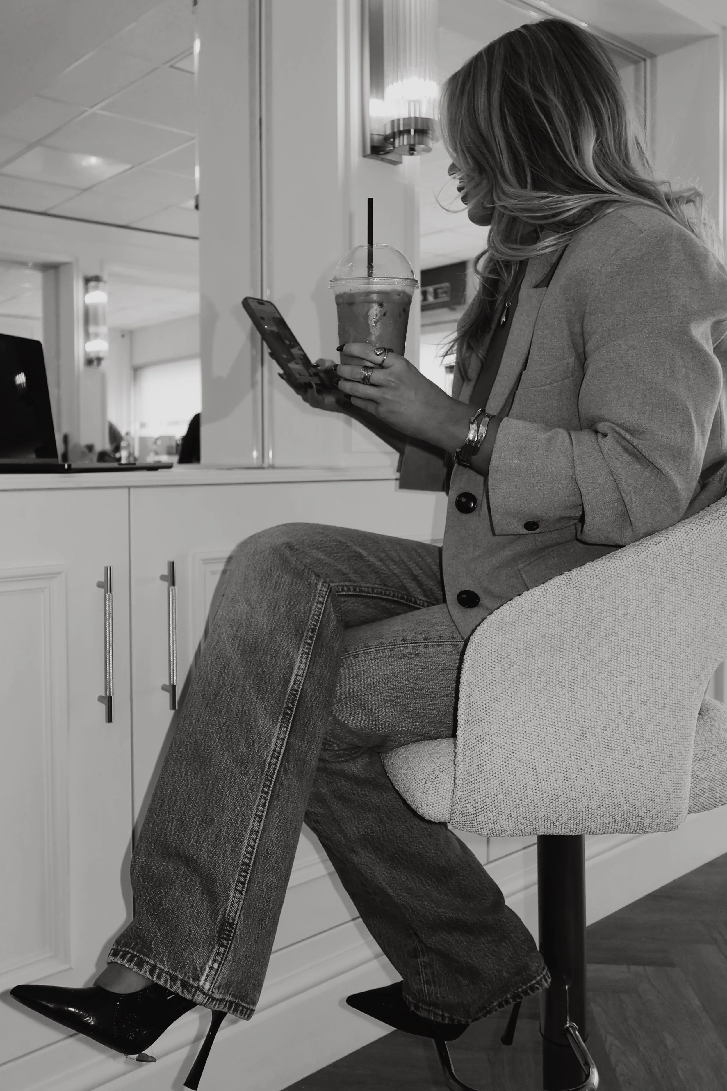 A woman with long hair sitting on a chair in a modern interior, wearing a blazer, jeans, and high heels, holding a large iced coffee and looking at her phone.