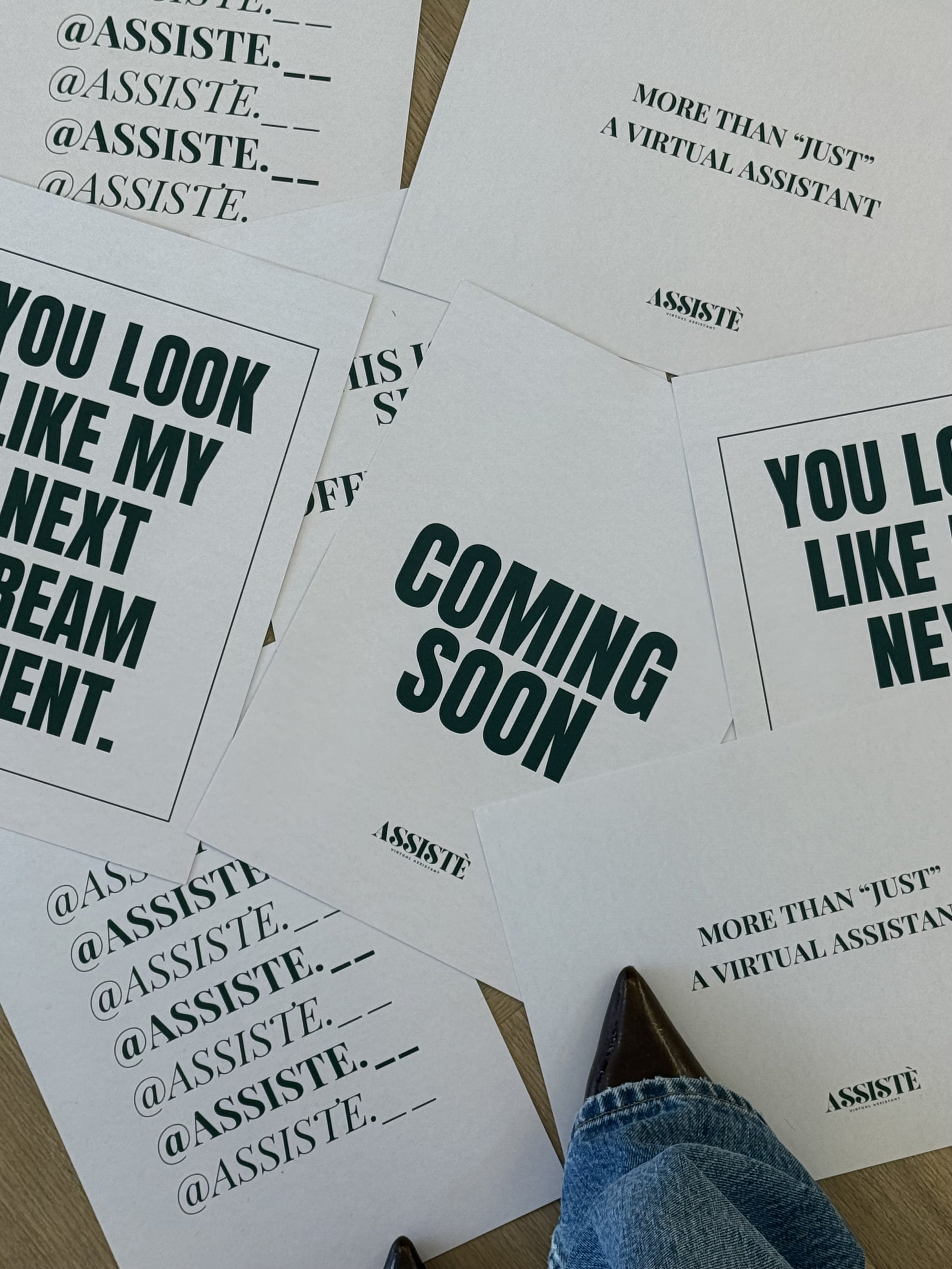 Various printed flyers and posters on a wooden surface, with messages like "COMING SOON," "YOU LOOK LIKE MY NEXT DREAM STUDENT," and details about a virtual assistant service called ASSISTÉ, including social media handles.