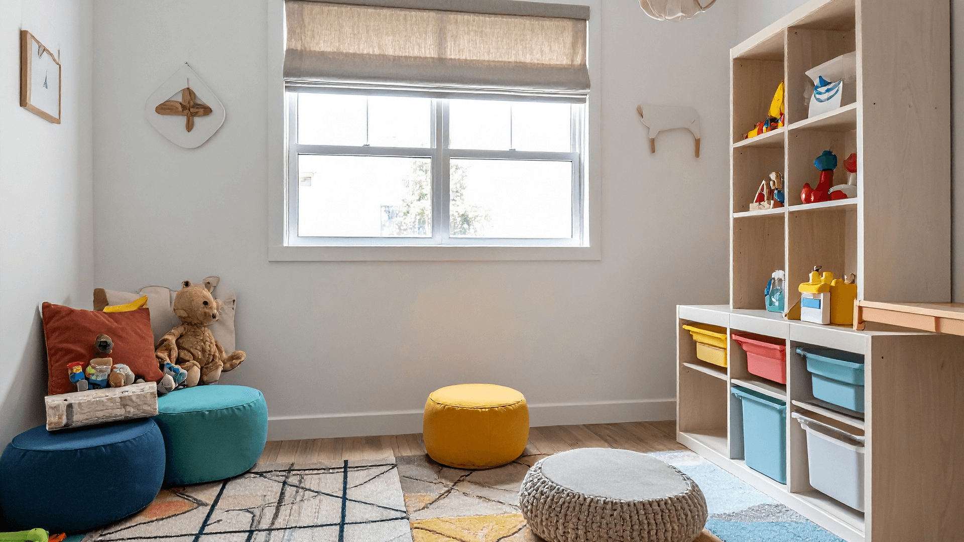 A cozy children's bedroom with a white wall, a window with a beige shade, and colorful plush toys and cushions. There is a storage shelf with various toys and bins, and a small seating area with soft poufs on a patterned rug.