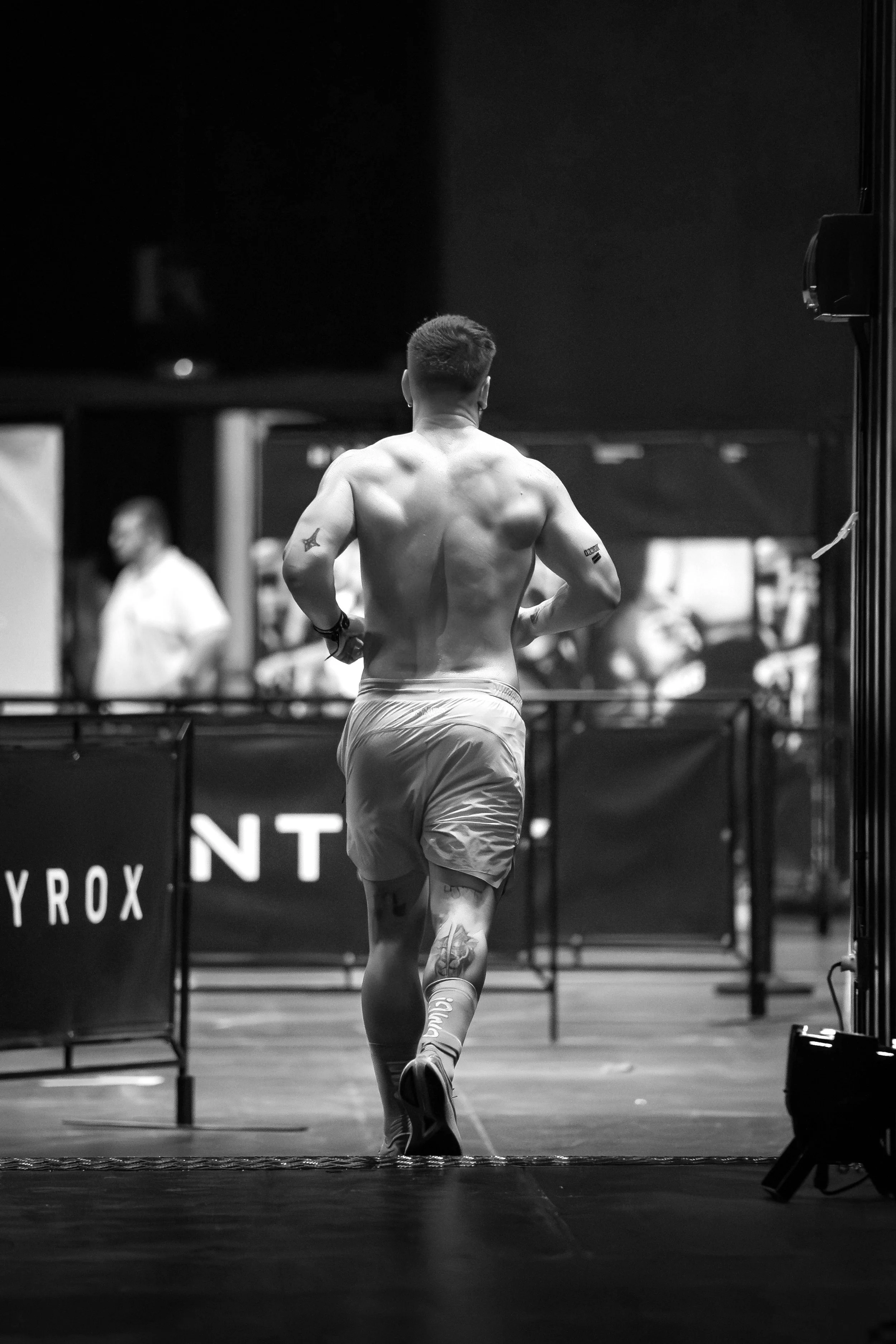Una persona sin camisa, vista desde atrás, corriendo en un evento de competencia. Lleva puesta ropa deportiva y tiene tatuajes en ambas piernas y en los brazos.
