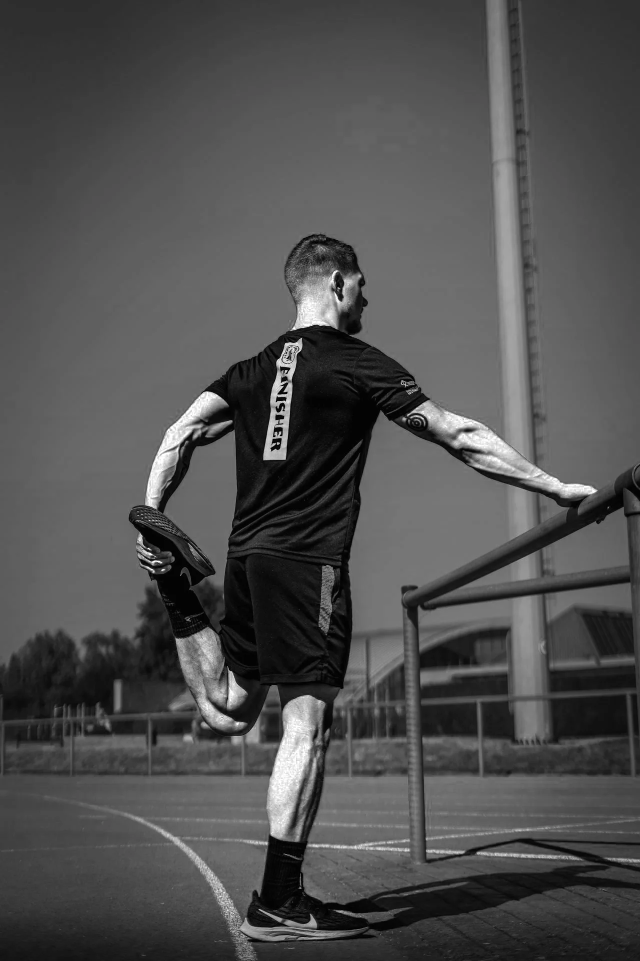 Hombre haciendo estiramiento en una cancha al aire libre, vista en ángulo lateral, en negro y blanco.