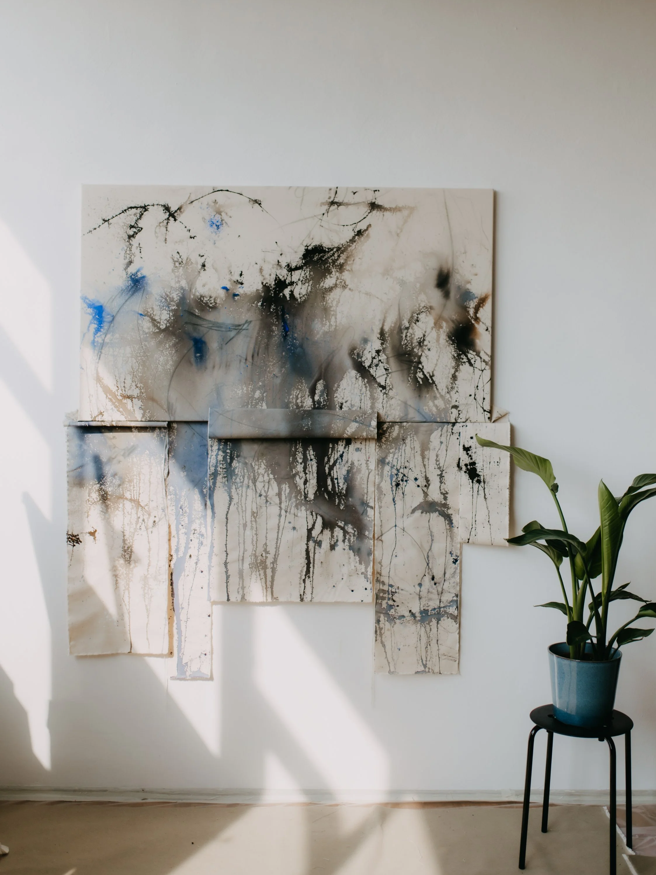 Abstract watercolor painting with drips and splatters in black, white, beige, and blue hues hanging on a white wall, with a green potted plant on a black stand nearby.