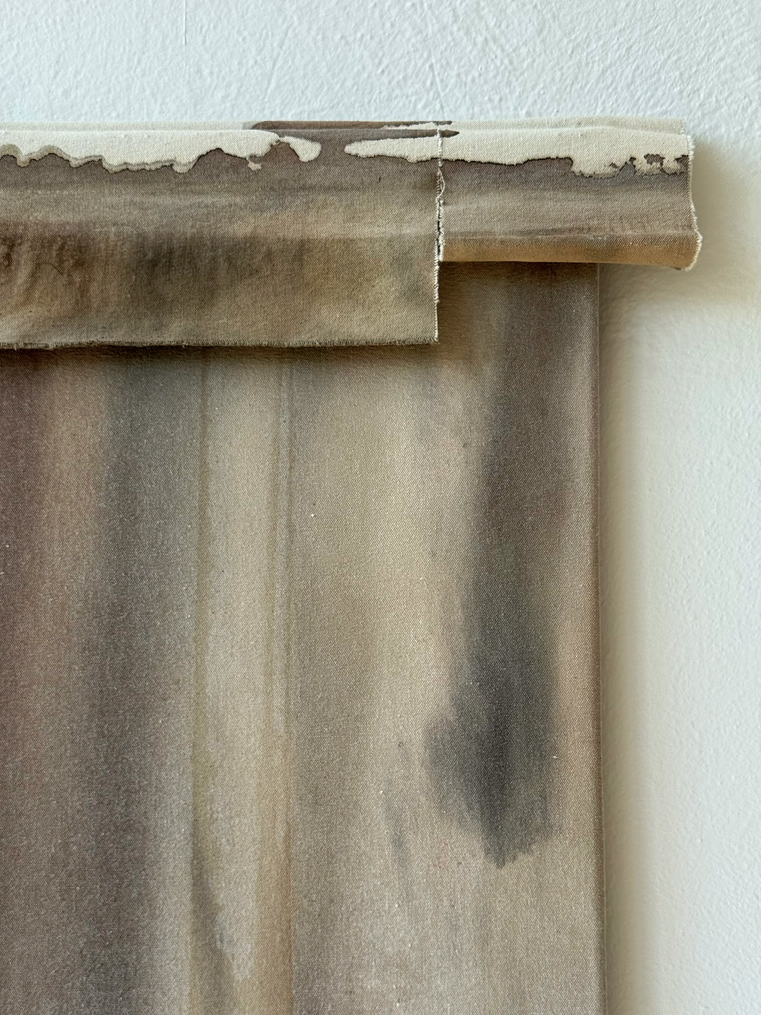 A beige and brown curtain partially pulled to the side, hanging against a white textured wall.