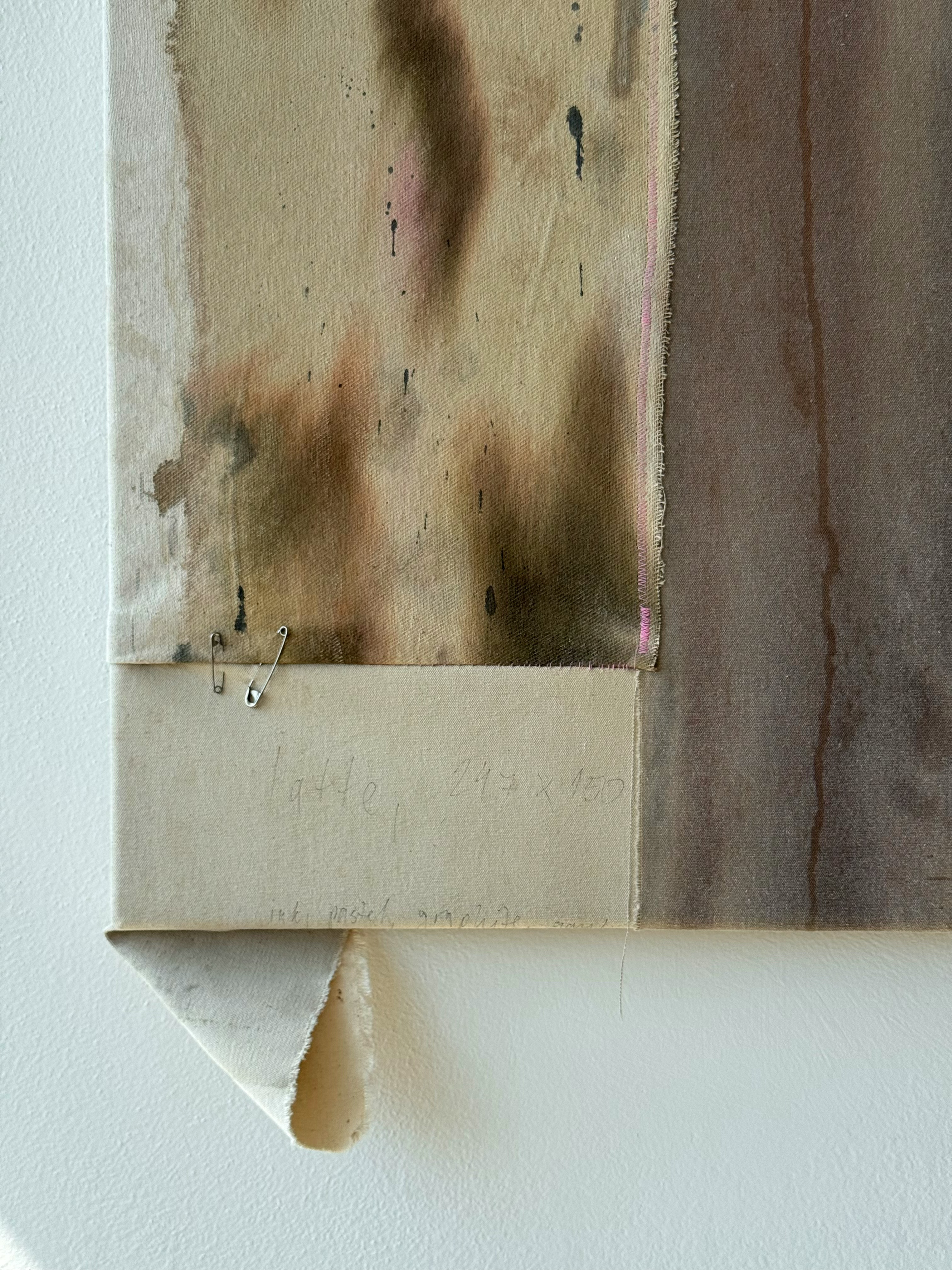 A mixed media art piece featuring a fabric with watercolor-like blotches of pink, brown, and black, including a textured fabric strip, two safety pins, and handwritten text on paper below the fabric.