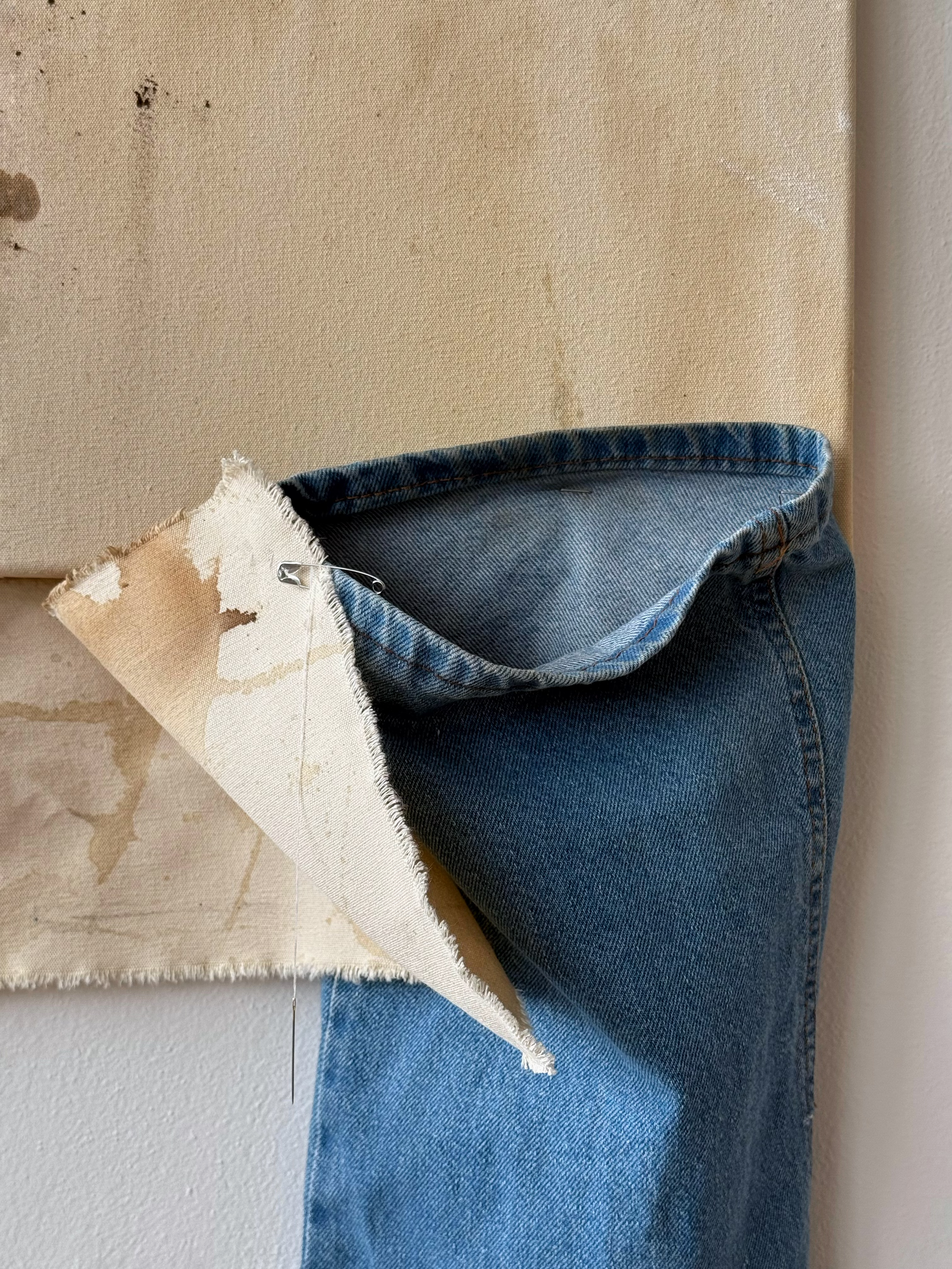Partially torn piece of fabric, possibly canvas or linen, with a safety pin attached, overlapping denim jeans hanging against a beige wall.