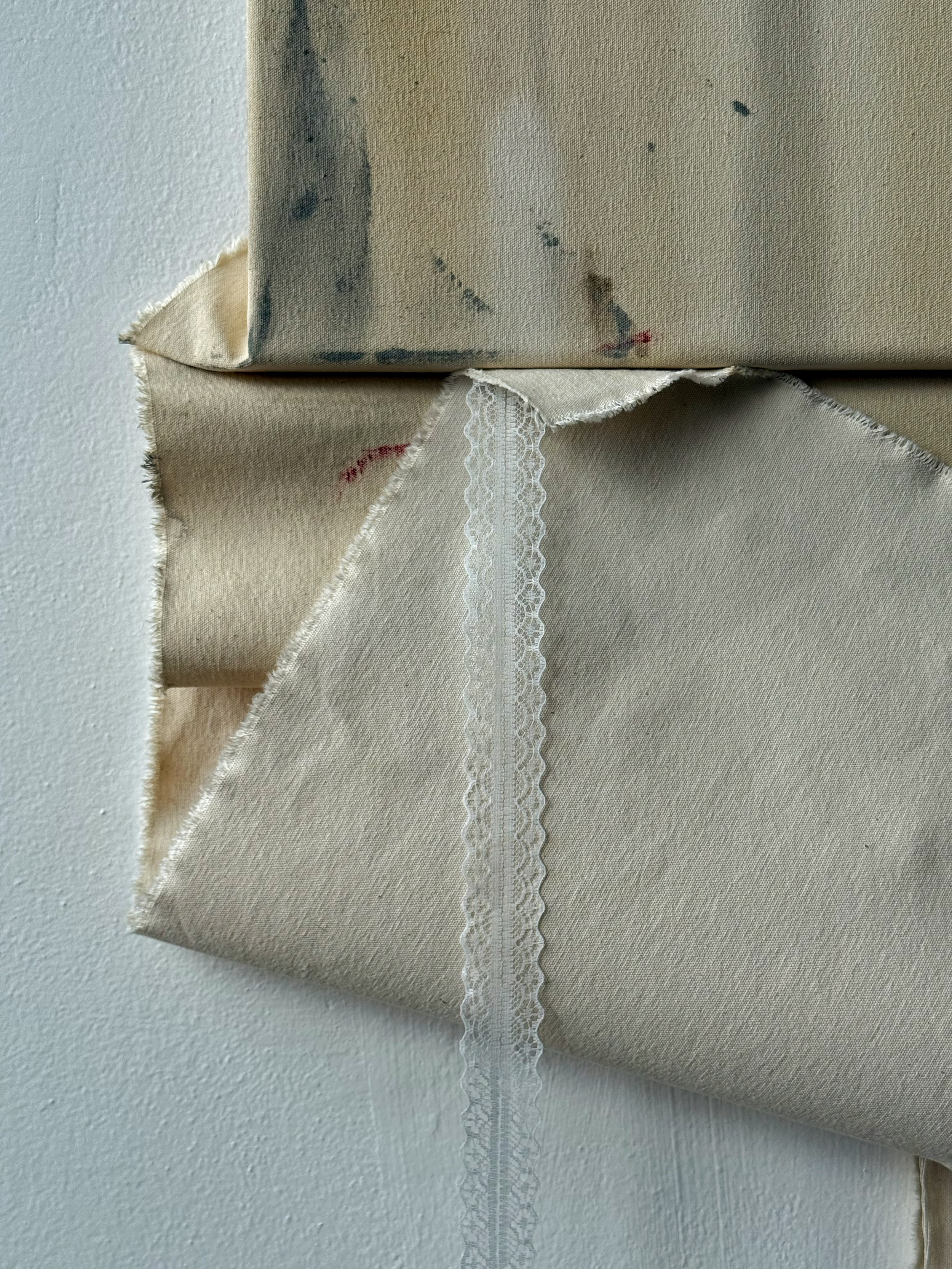 Close-up of beige fabric with edges frayed and lace trim, hanging against a white textured wall.
