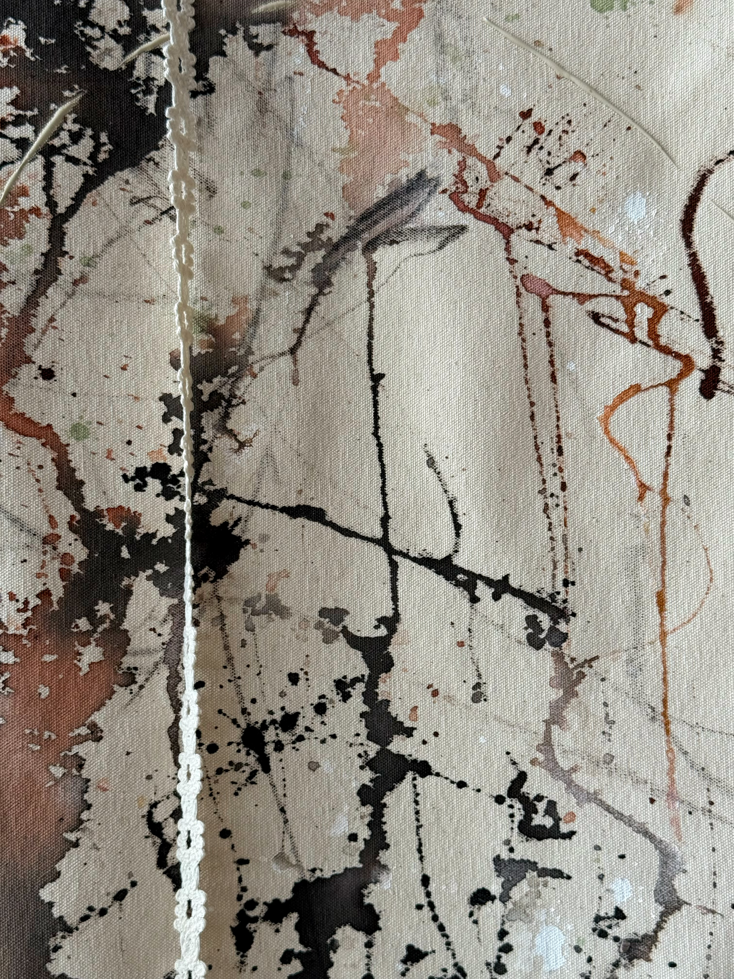 Close-up of abstract splattered paint on canvas, with black, brown, and beige tones, and textured vertical white thread or cord running down the middle.