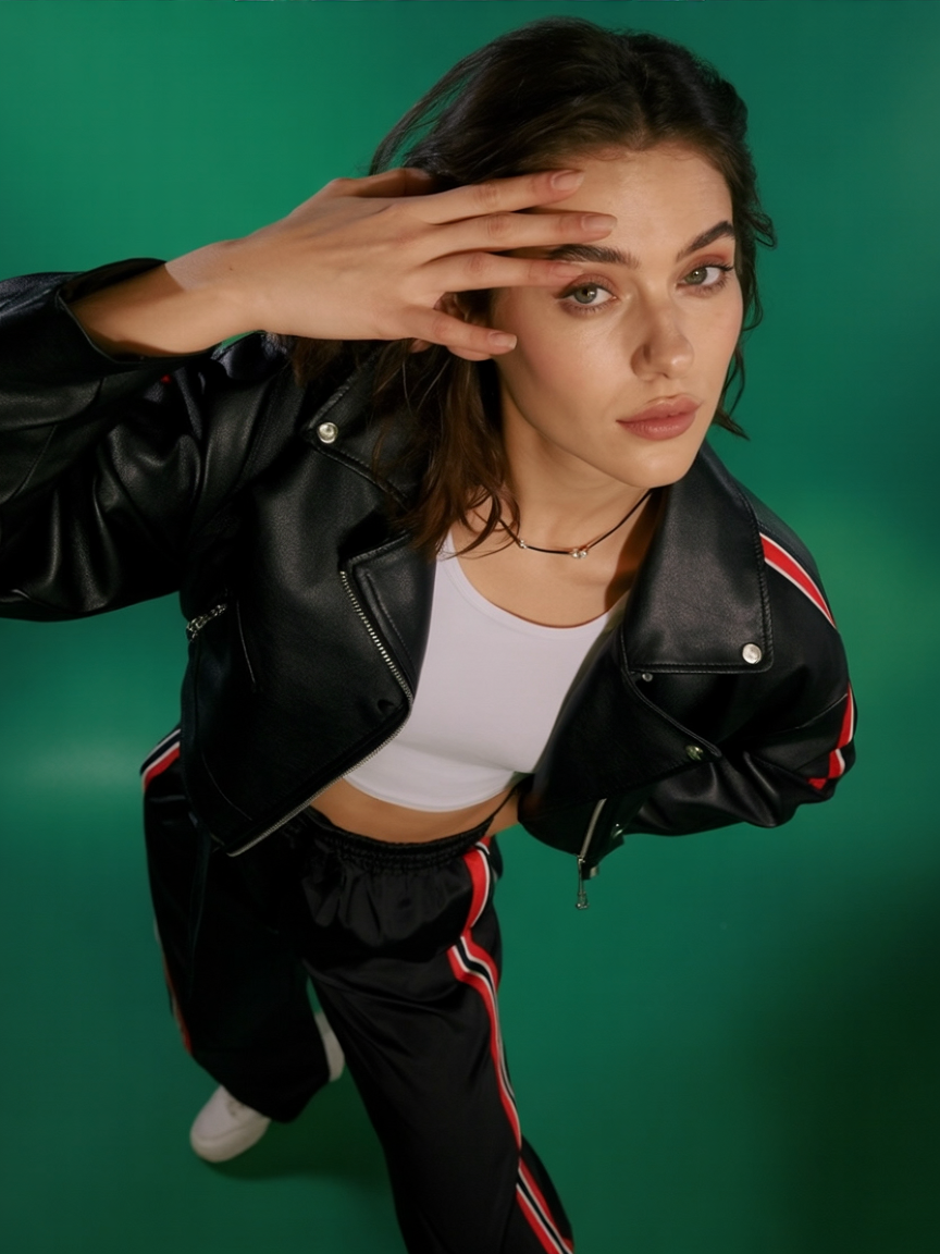 A woman wearing a black leather jacket, white crop top, and black pants with red and white stripes, adjusts her hair with her hand while looking at the camera against a green background.