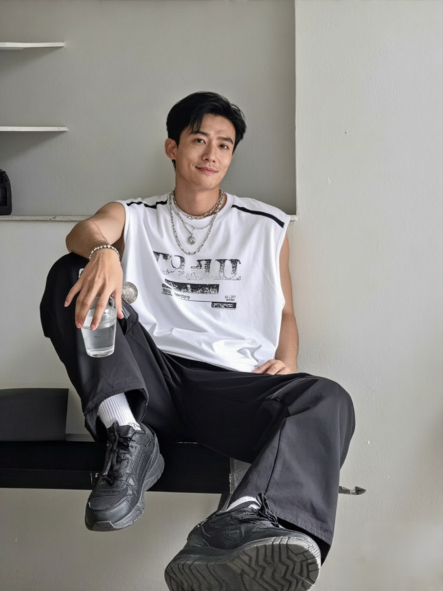 A young man with black hair sitting on a black bench against a white wall, holding a glass of water, wearing a white sleeveless graphic T-shirt, black pants, and black sneakers, accessorized with layered silver necklaces, earrings, and bracelets, smiling softly at the camera.