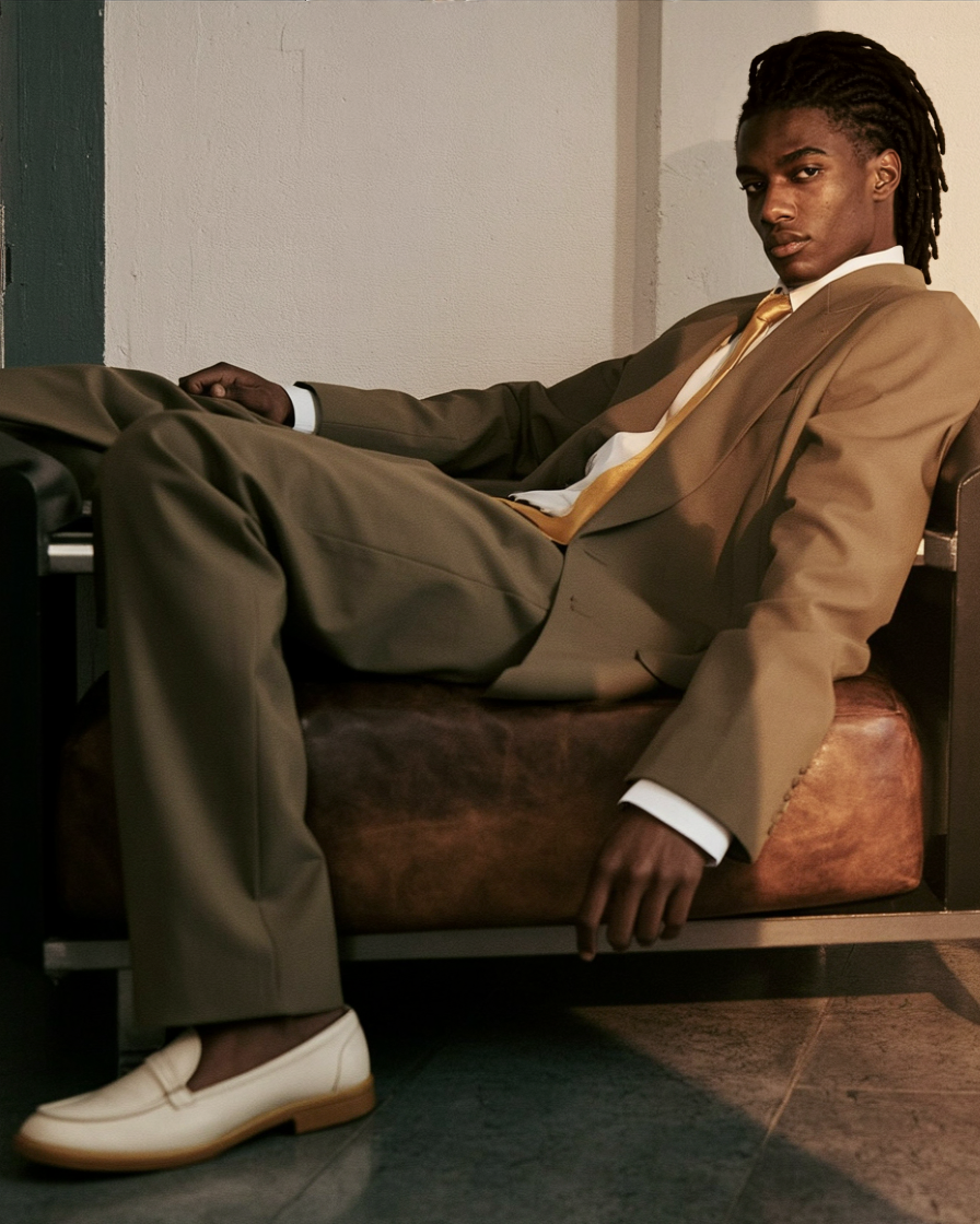 A young man with dreadlocks sitting on a leather couch wearing a tan suit and white shoes, looking at the camera.