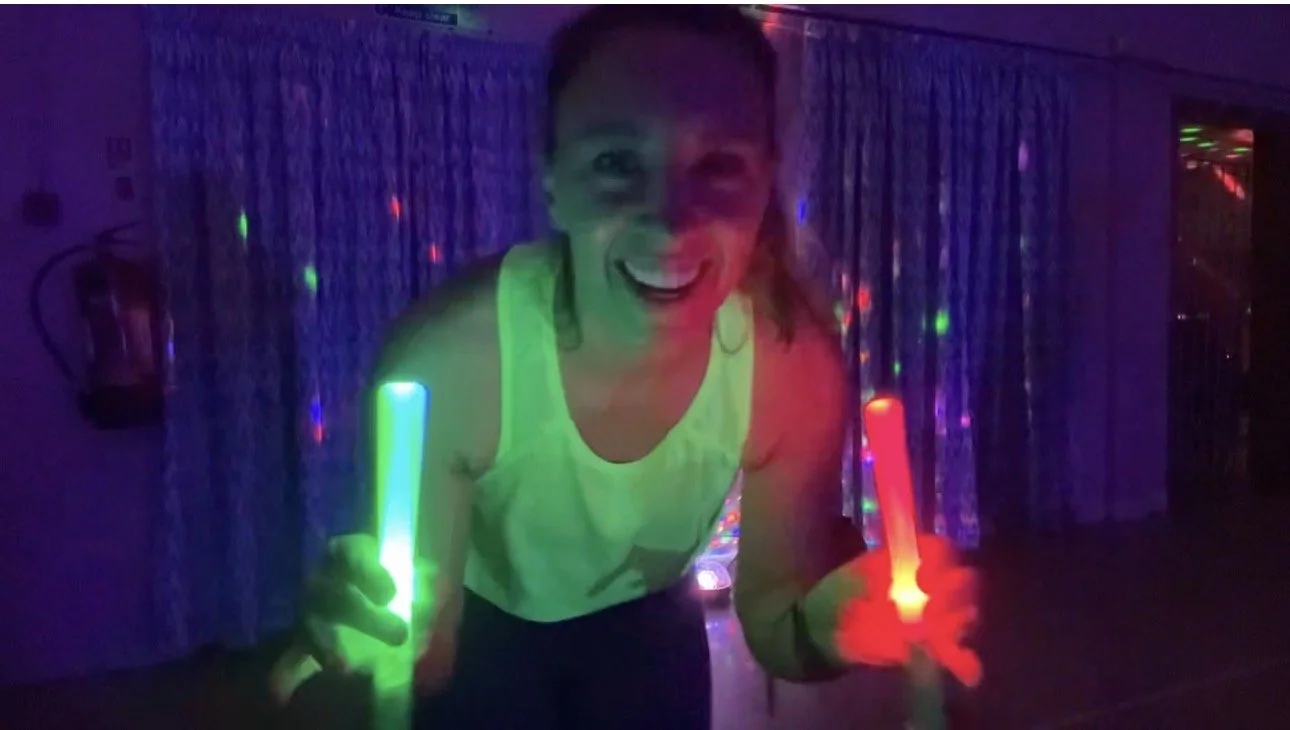 dark room with disco lights, woman in bright green vest top with glow sticks, smiling to camera