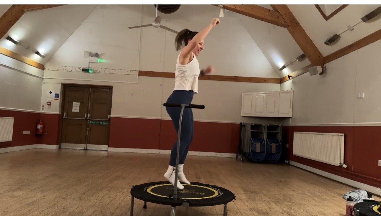 woman jumping on mini fitness trampoline, arm in air, smile on face, side facing