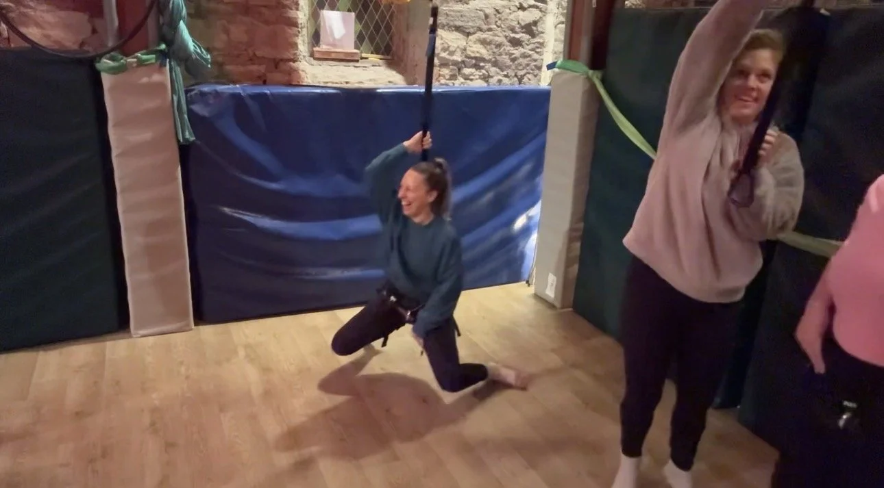 bungee fitness class, instructor setting up lady on kit, another female haning from celing by a bungee cord and harness, she is laughing.