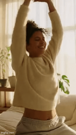 A woman stretching and smiling in a bright, cozy room with sunlight, plants, and a bed.