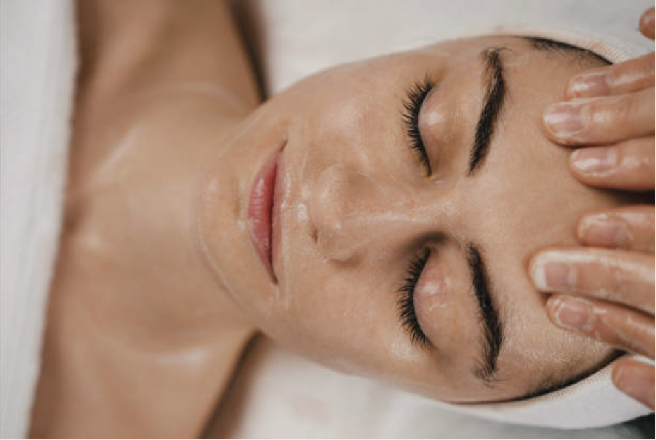 A woman receives a facial massage at a spa, lying peacefully with her eyes closed while a therapist applies gentle pressure on her forehead.