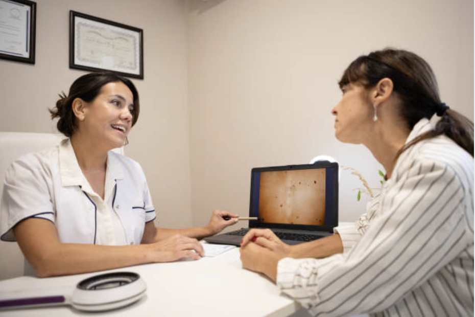Two women in a medical consultation: one is a doctor or healthcare professional holding a pen, and the other is a patient talking. They are seated at a table with a laptop, and behind them are framed certificates on the wall.