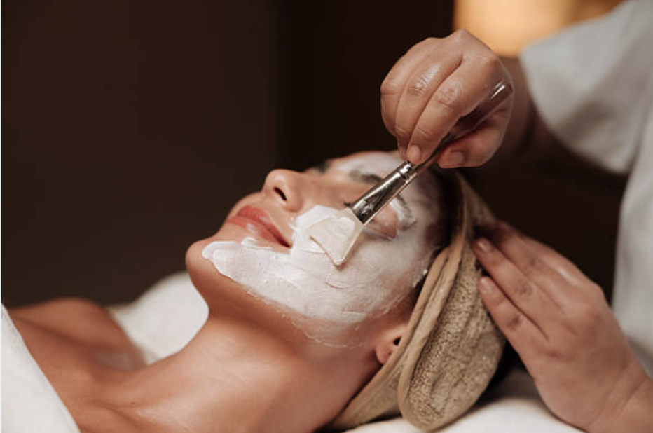 A woman receiving a facial treatment with a white cream mask being applied by a professional therapist at a spa or skincare clinic.