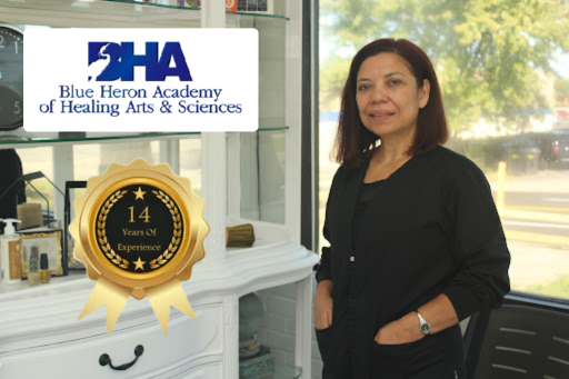 A woman standing inside a building next to a display with a sign that says 'Blue Heron Academy of Healing Arts & Sciences'. The woman is wearing a black jacket and has shoulder-length dark hair, with her hands in her pockets. There is a window behind her showing trees and parked cars outside.