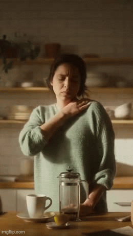 A woman standing in a kitchen, touching her neck, with a French press and a mug on the counter in front of her.