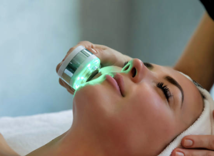 A woman receives a skincare treatment with a device emitting green light on her chin and nose.