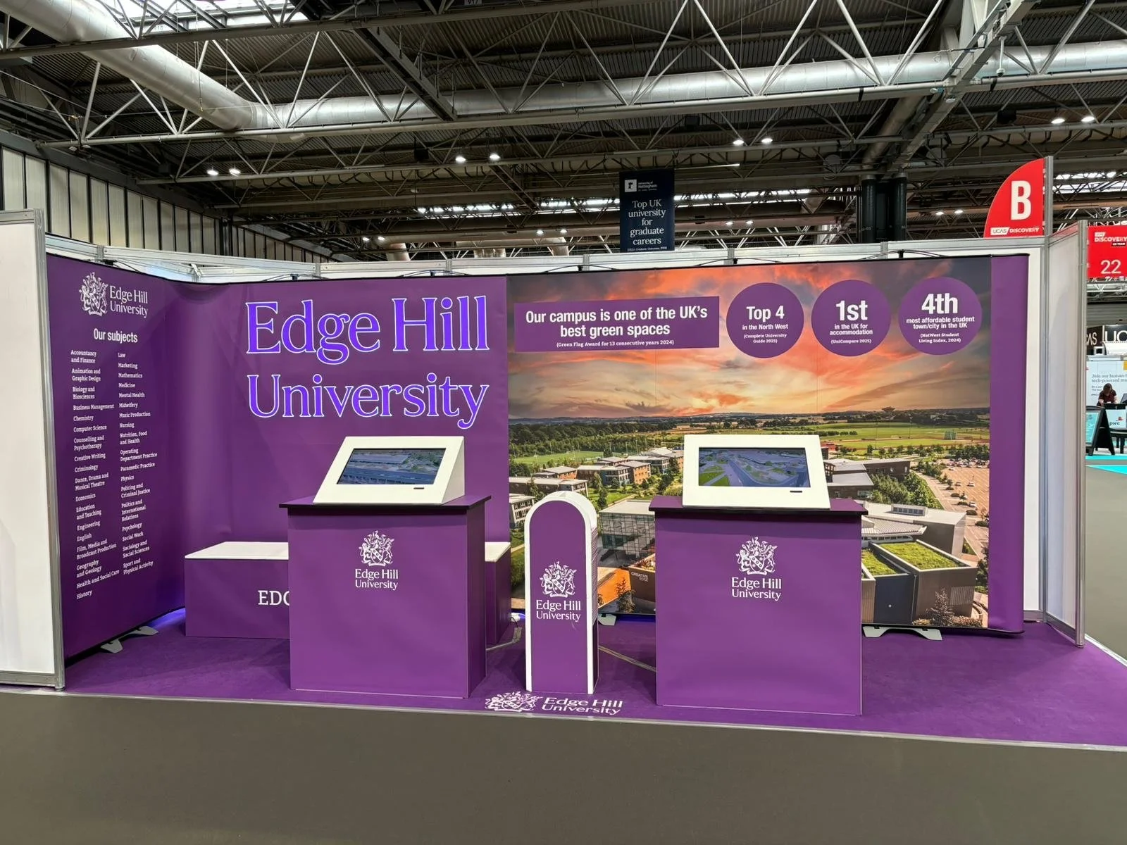 A booth for Edge Hill University at an educational fair with purple background, two tablet displays, and promotional signage highlighting the university's ranking and campus green spaces.