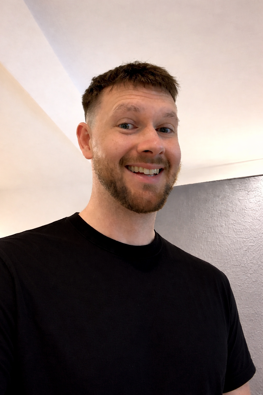 A smiling man with short brown hair, a beard, and blue eyes wearing a black T-shirt, standing indoors against a beige and gray wall.