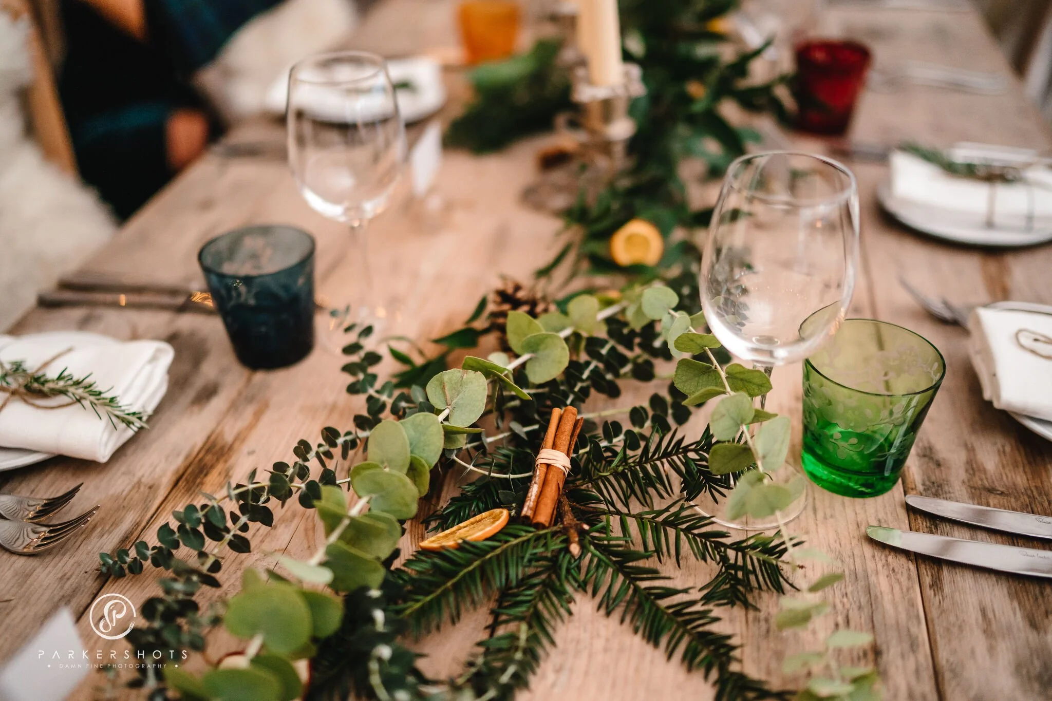 A wooden table set for a meal with glassware, napkins, and silverware, decorated with greenery and citrus slices.