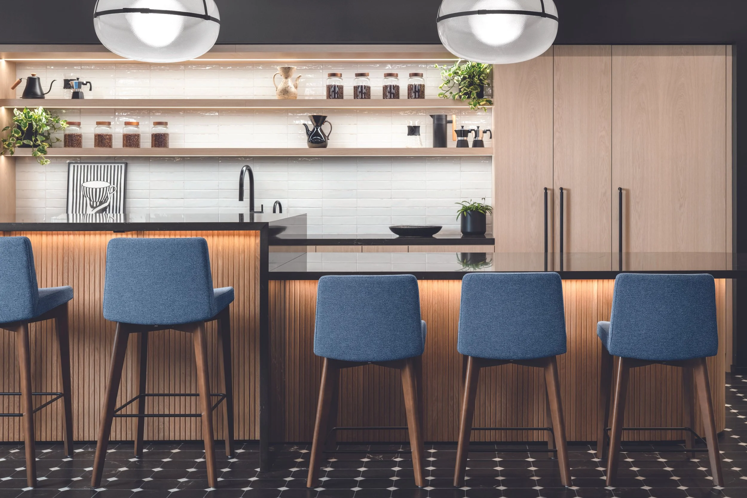 Modern kitchen with black countertops, wooden cabinets, white subway tile backsplash, and blue upholstered barstools at a black and wooden bar counter. Open shelves with decorative jars and plants. Overhead hanging pendant lights.