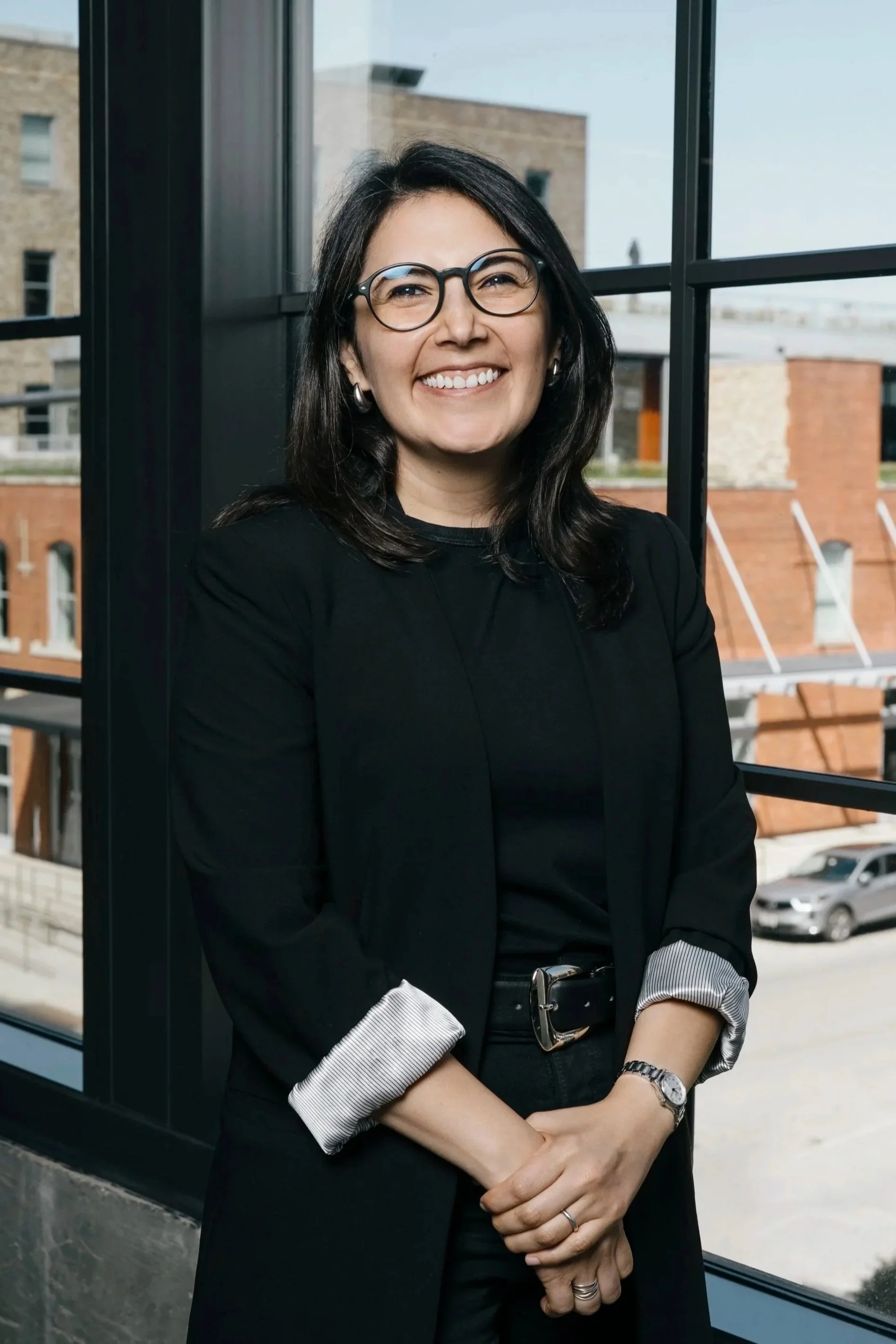 A woman with dark hair and glasses smiling in front of a window, wearing a black blazer with rolled-up sleeves, showing a blueprint underneath.