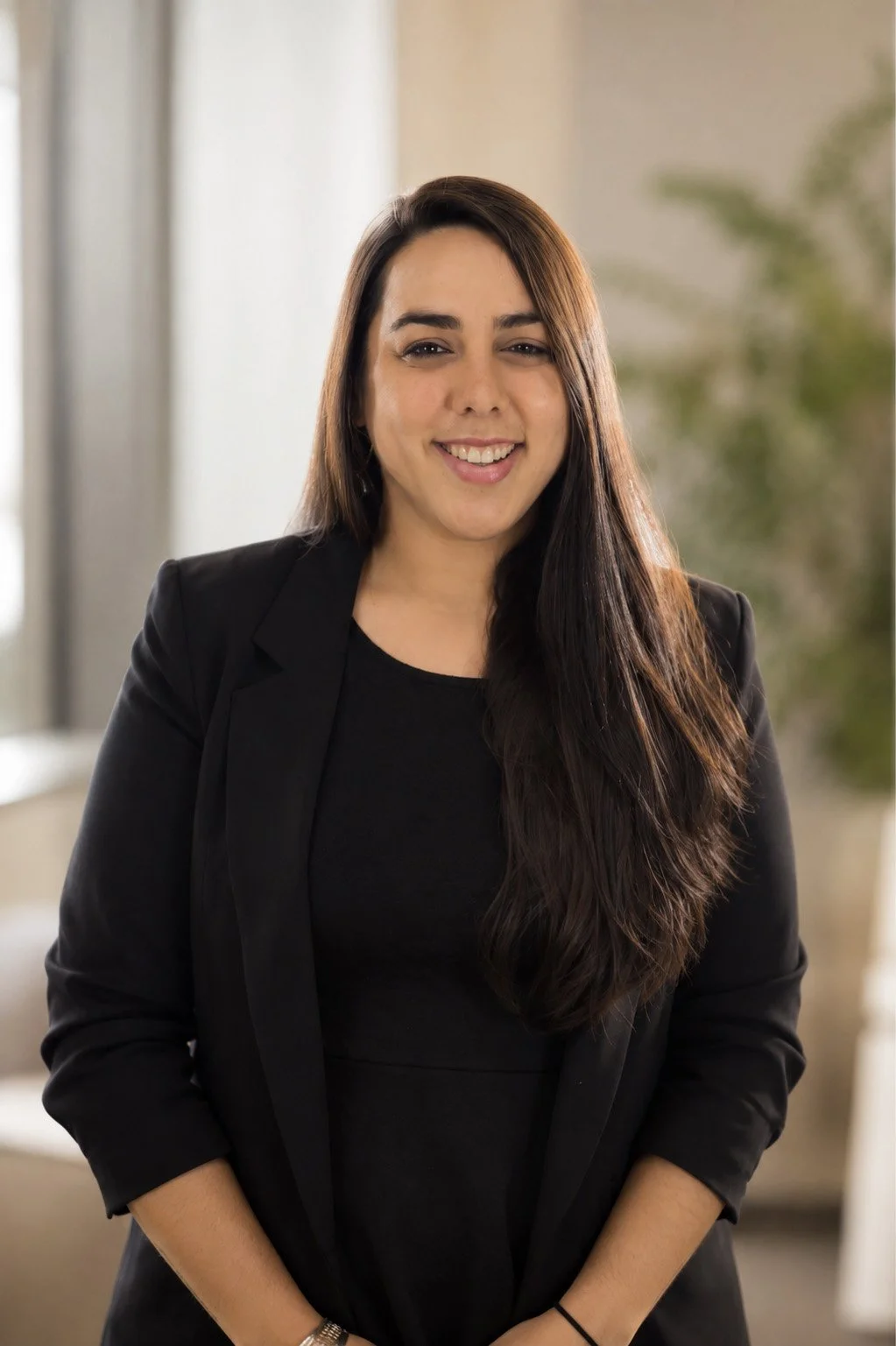 A young woman with long dark hair smiling and wearing a black blazer over a black shirt, standing indoors with a blurred background.