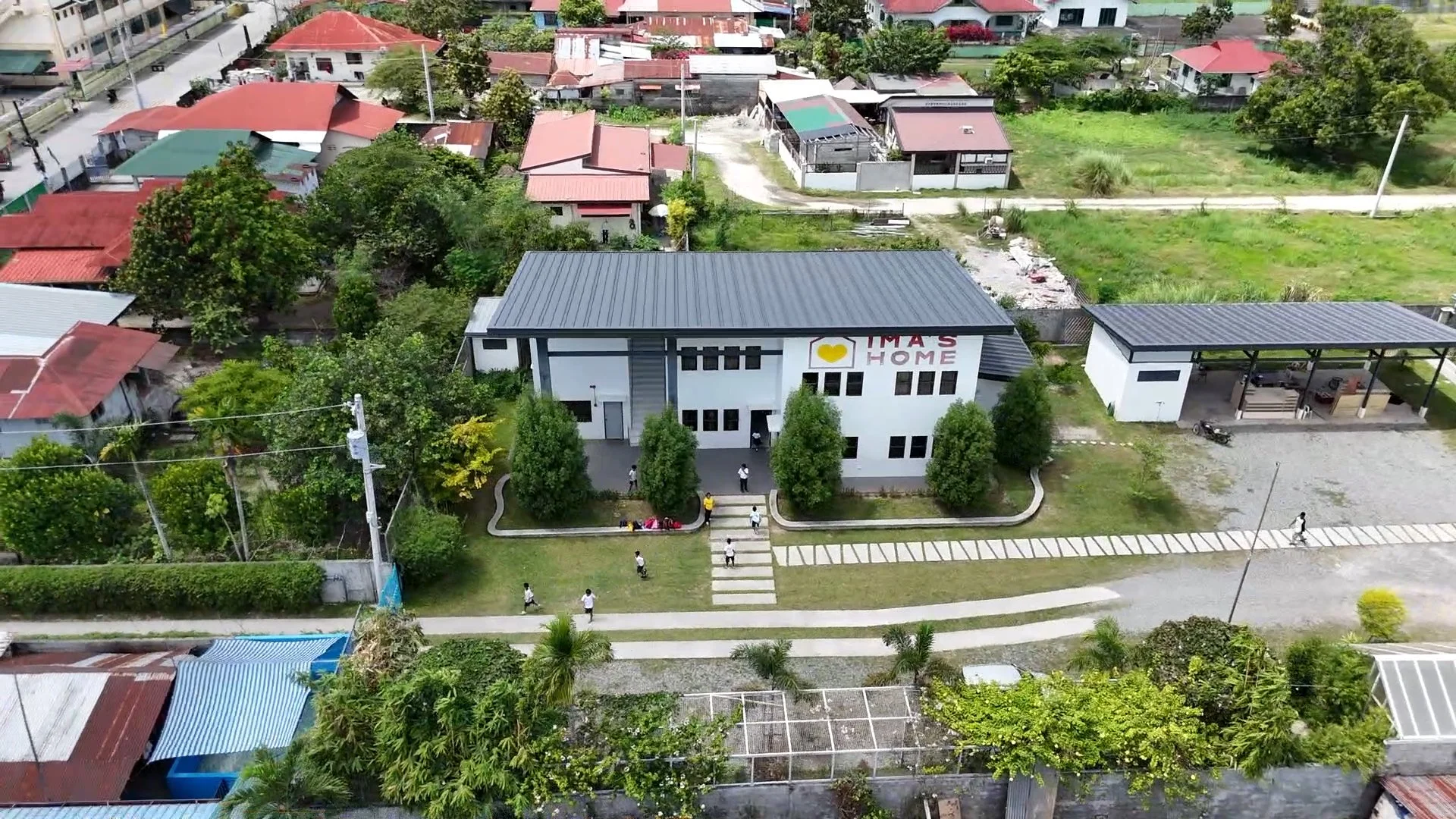An aerial view of a school building with a sign that reads 'I'M AT'S HOME,' surrounded by trees, students walking on the campus, neighboring houses, and green fields.