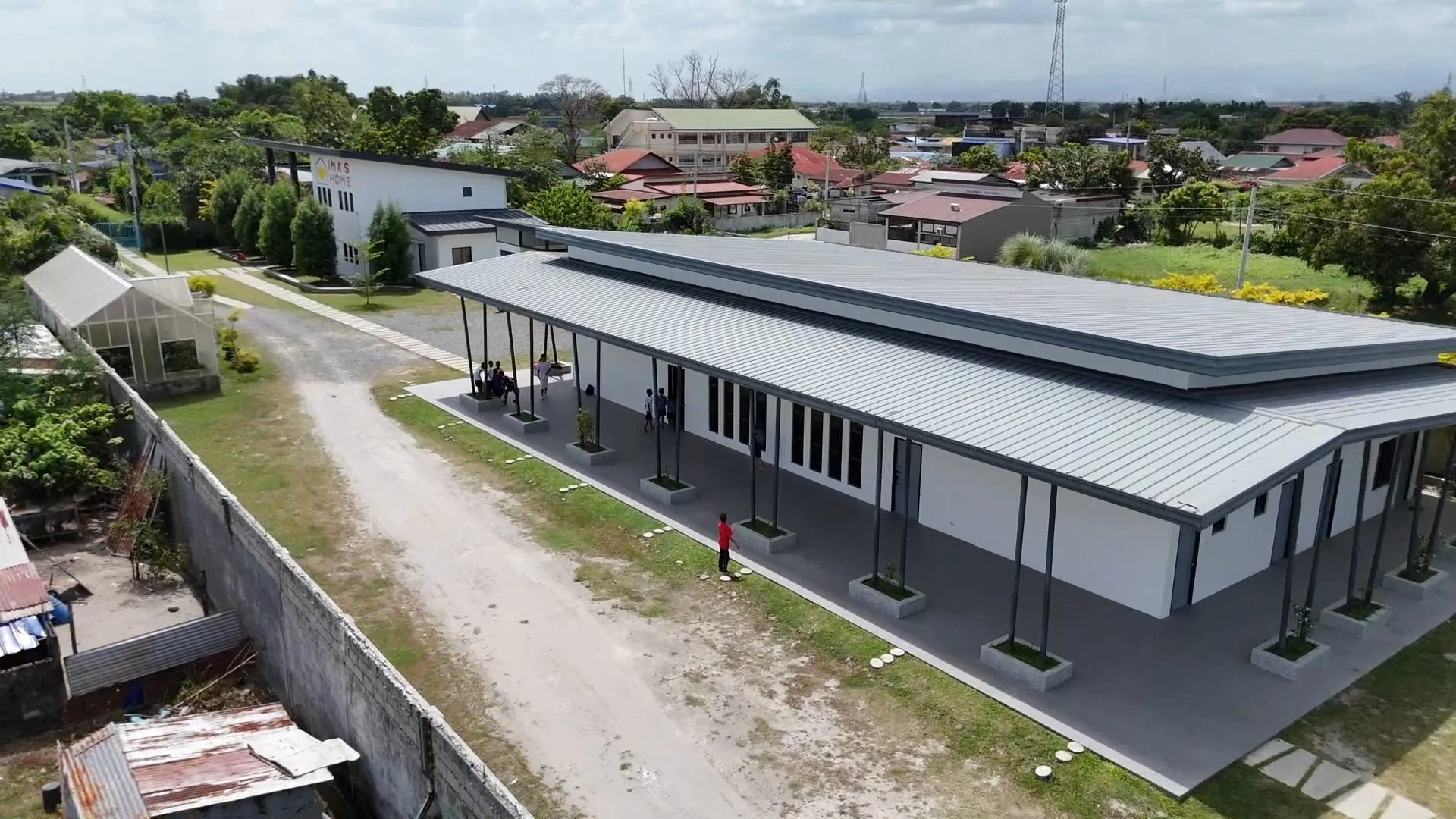 Aerial view of a modern building with a metal roof and tall support columns, surrounded by a landscape with trees, gravel paths, and nearby residential houses.