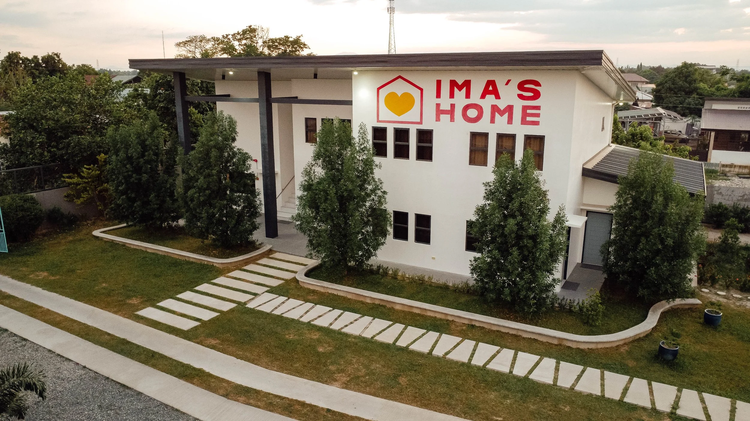 A modern two-story house with a large sign that says "Ima's Home", featuring a yellow heart inside a red outline of a house. The house has white walls, several small windows, and is surrounded by a well-maintained lawn with trees and a curved pathway.