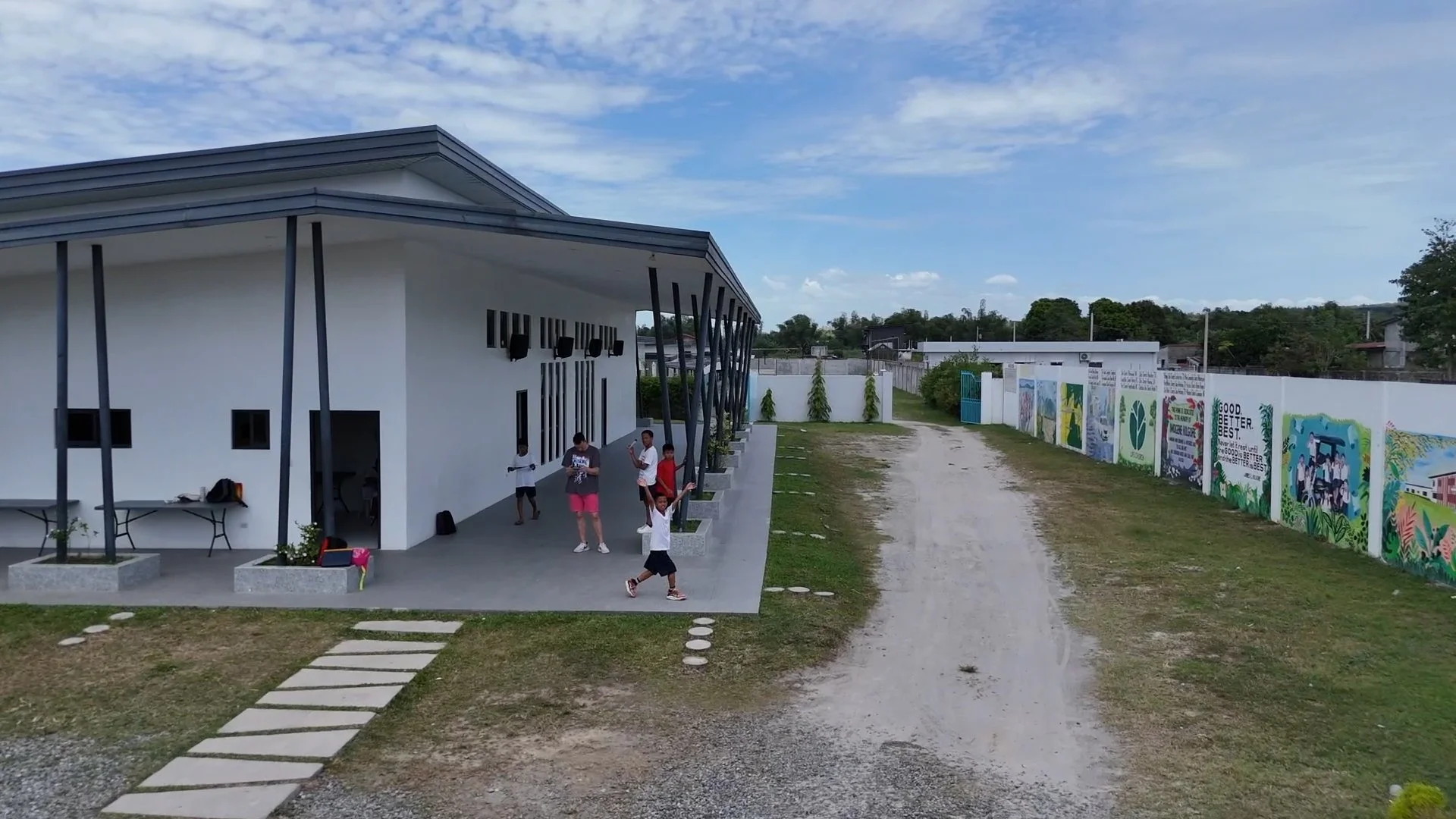 A group of children playing outside a modern white building with black window frames and vertical support beams, next to a colorful mural on a white wall, with a gravel road and grassy area in the surroundings under a partly cloudy sky.