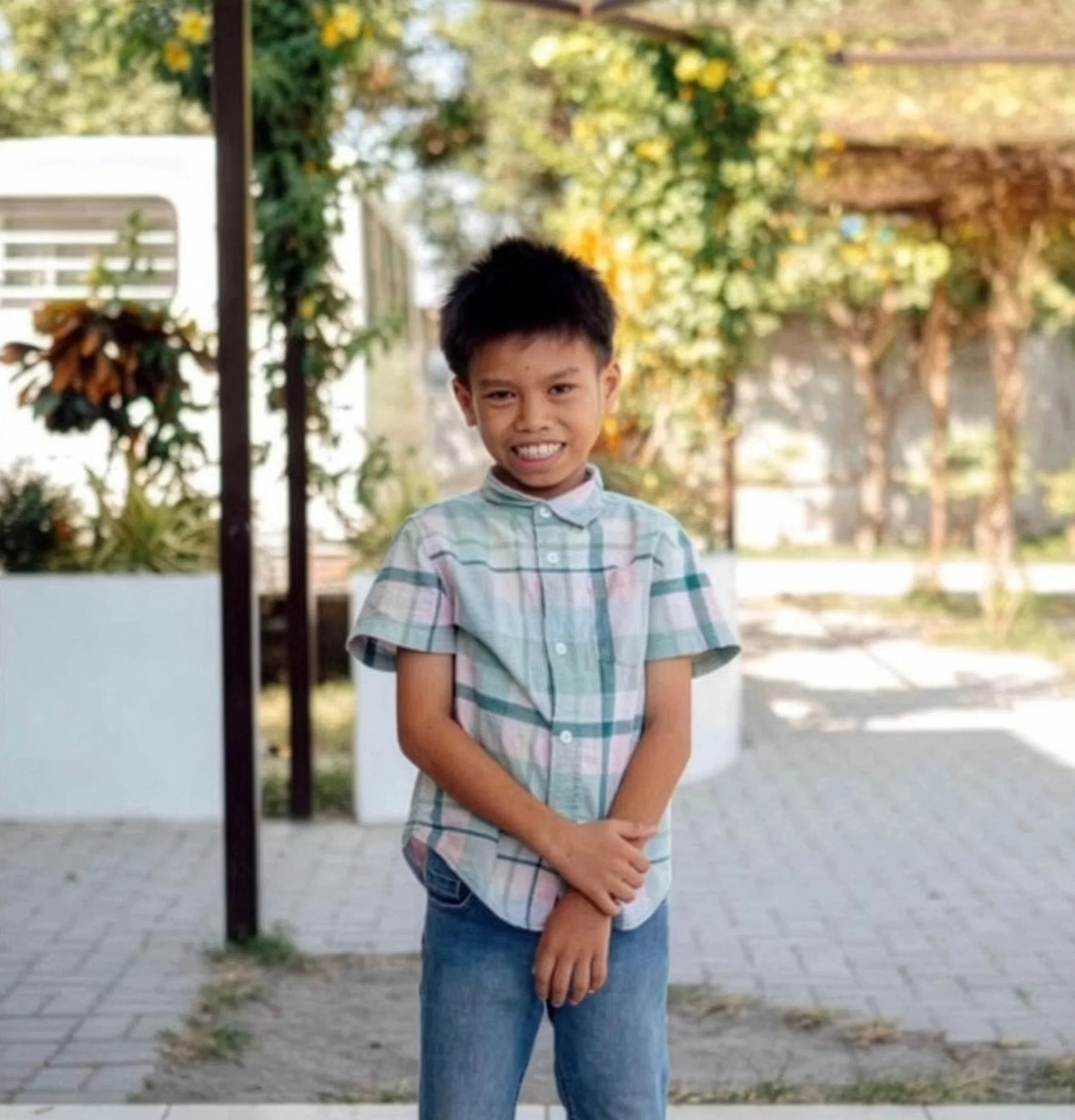A young boy standing outdoors on a paved area, smiling and holding his hands in front of him. Background features trees with green and yellow leaves and some modern structures.