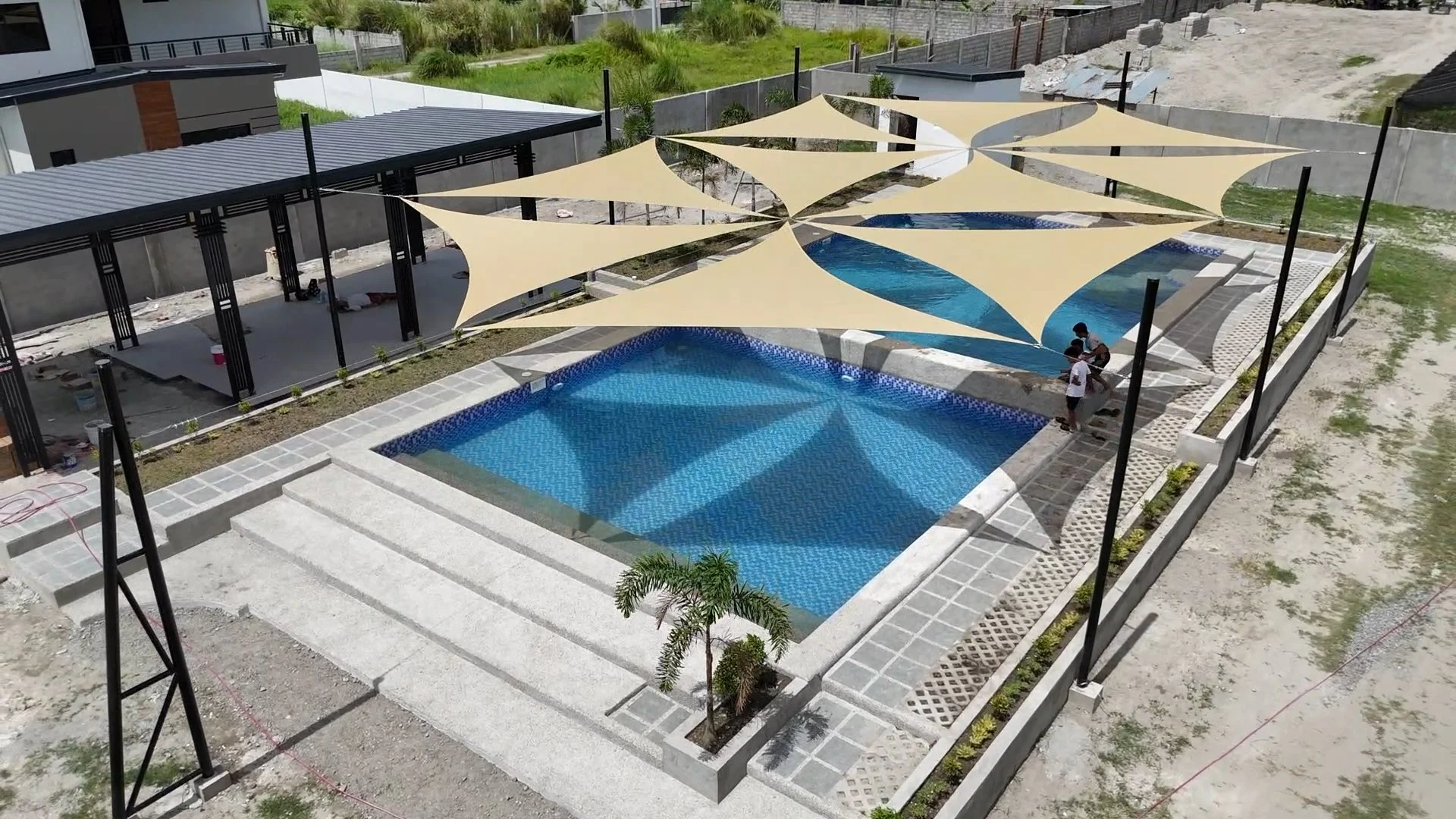 Aerial view of a swimming pool with three beige shade sails overhead, in a construction site with some workers, surrounded by a concrete walkway and a fence, with upcoming landscaping.
