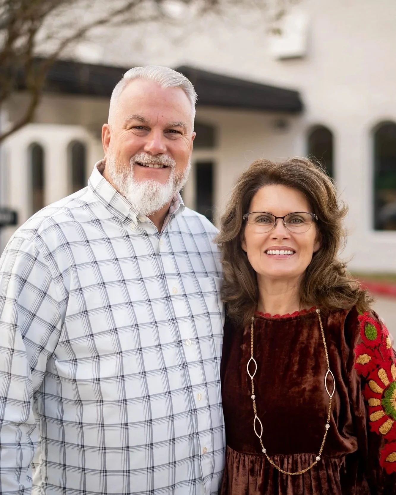 A smiling elderly couple standing outside a house, with the man wearing a white plaid shirt and the woman in a velvety brown top with embroidered sleeves and glasses.
