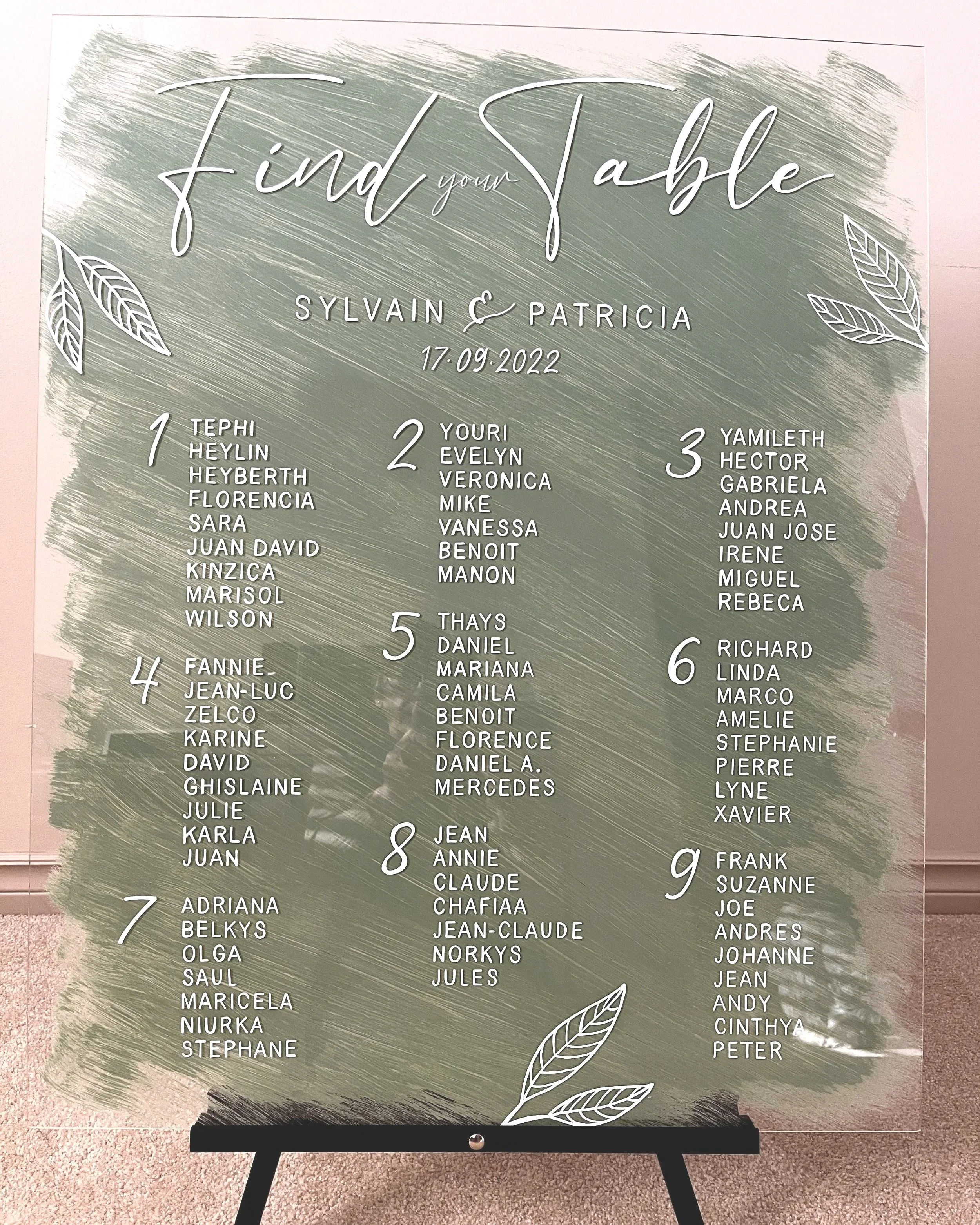Seating chart with the title 'Find your Table' for Sylvain and Patricia wedding on September 17, 2022. The chart lists nine tables with guest names grouped under each table number, decorated with white leaf illustrations on a green brushstroke background.