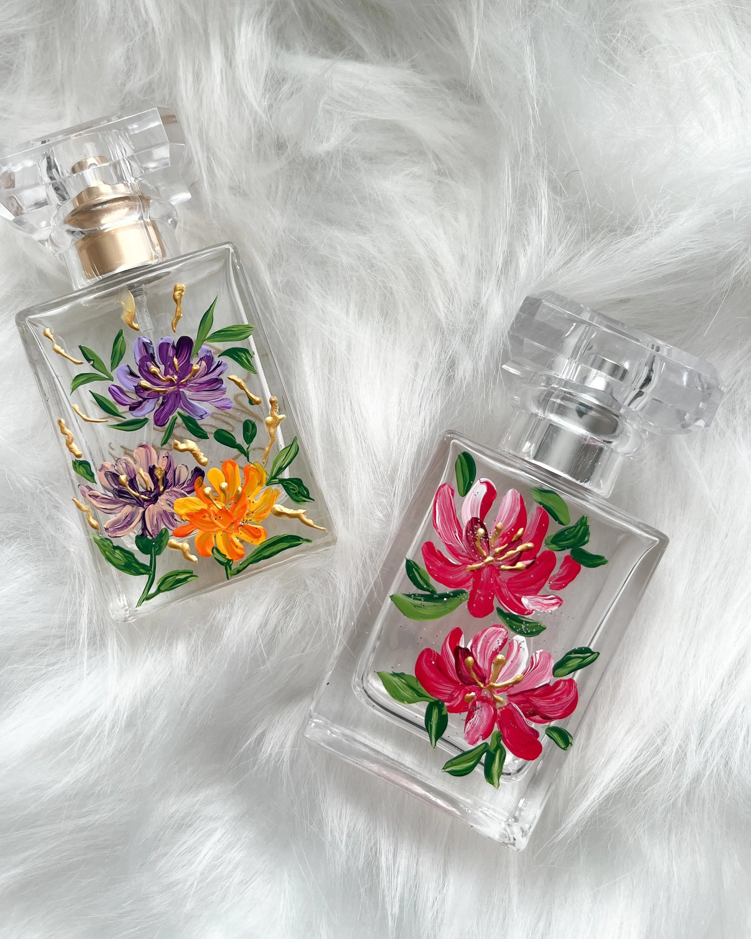 Two perfume bottles with hand-painted floral designs, one featuring purple and orange flowers and the other pink flowers, placed on white fur background.