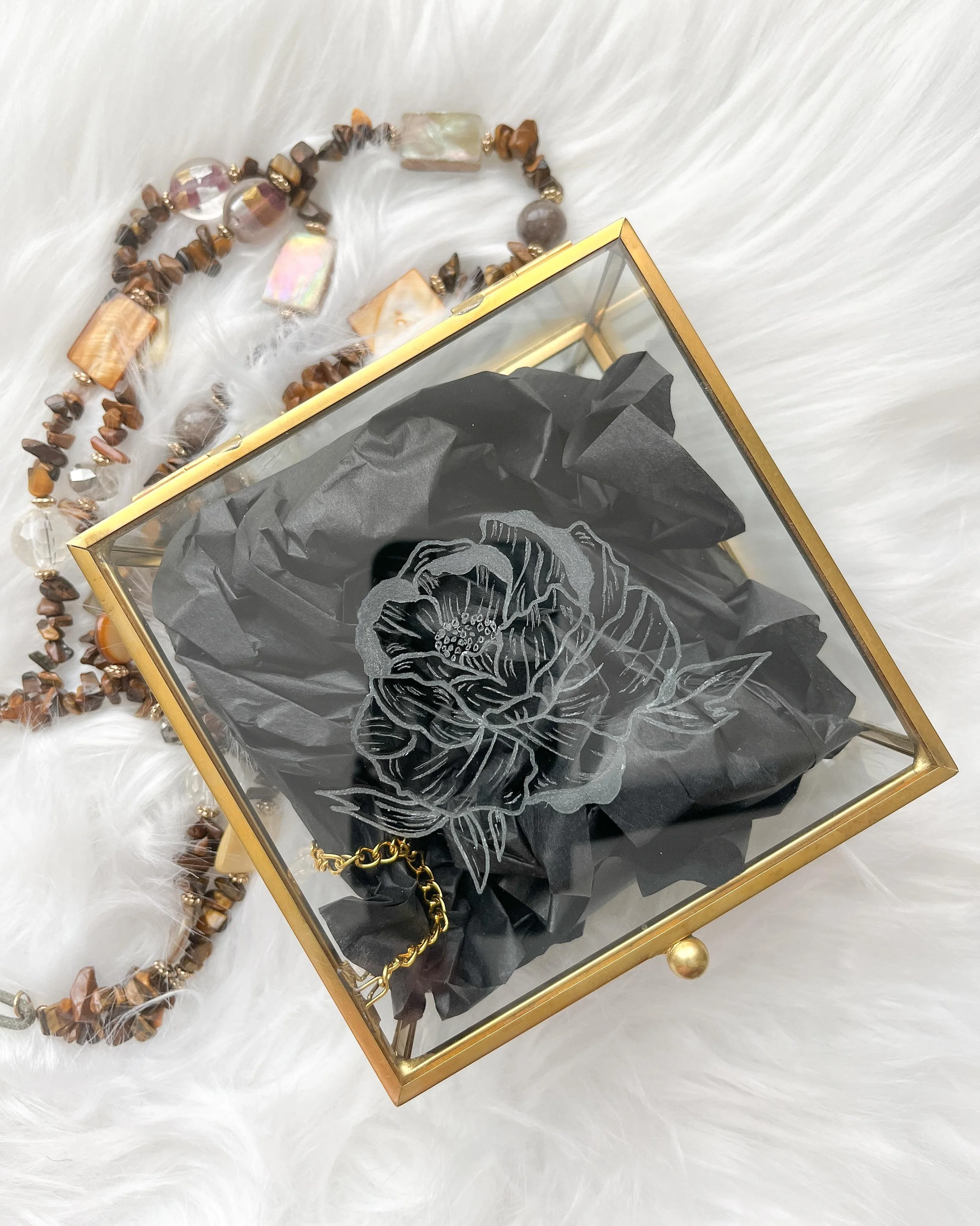 A glass box with a gold frame containing crumpled black tissue paper and a gold chain, placed on a white furry surface. A beaded necklace made of various brown, beige, and iridescent beads is nearby.