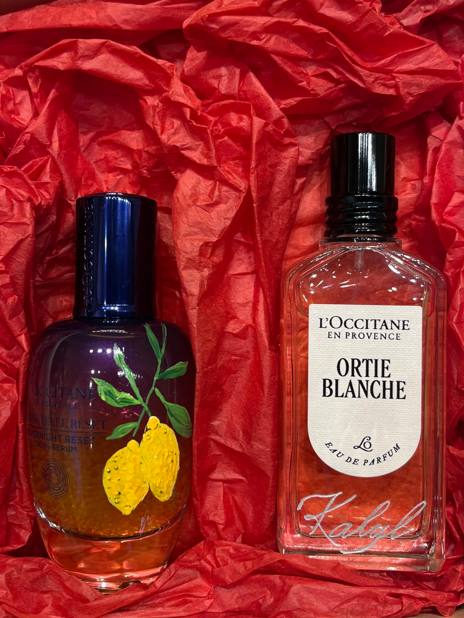A gift box containing a bottle of L'Occitane en Provence perfume and a serum, wrapped in red tissue paper.