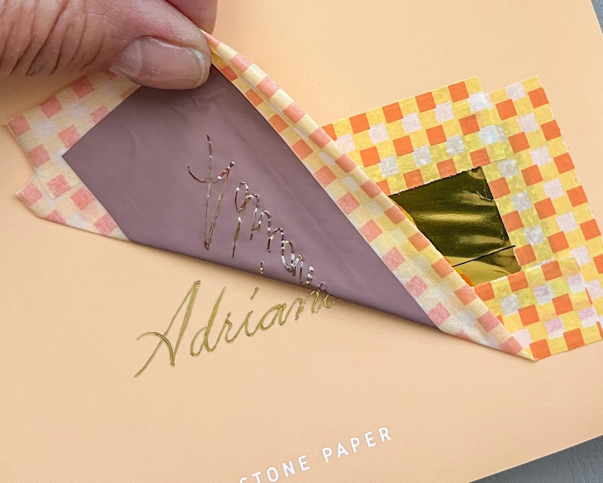 A person peeling back the corner of a pink and orange checkered paper revealing a gold foil behind. The paper is on top of an embossed cardstock that reads 'Adriana' with part of the word 'Thank' visible.