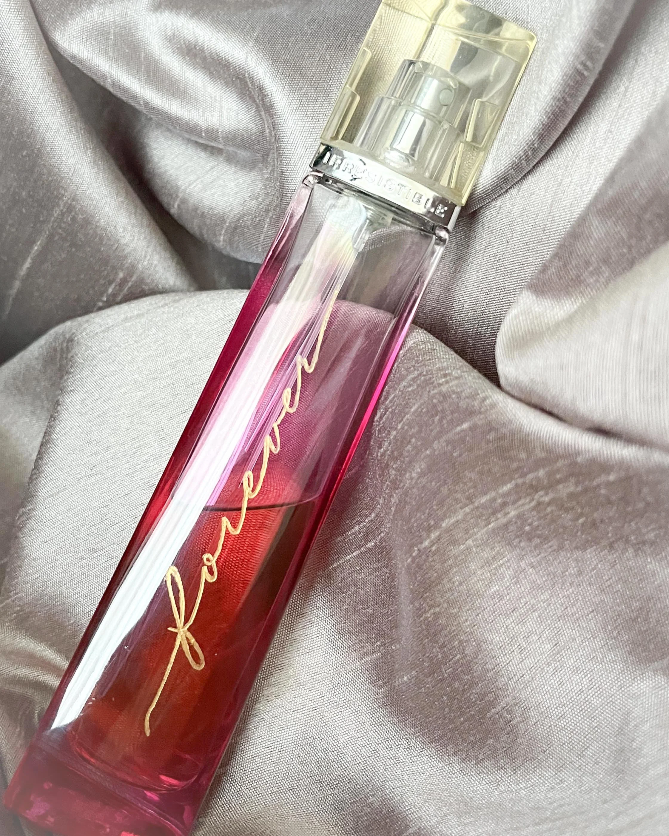 A transparent perfume bottle with pink liquid and gold script reading 'famous' rests on metallic satin fabric.