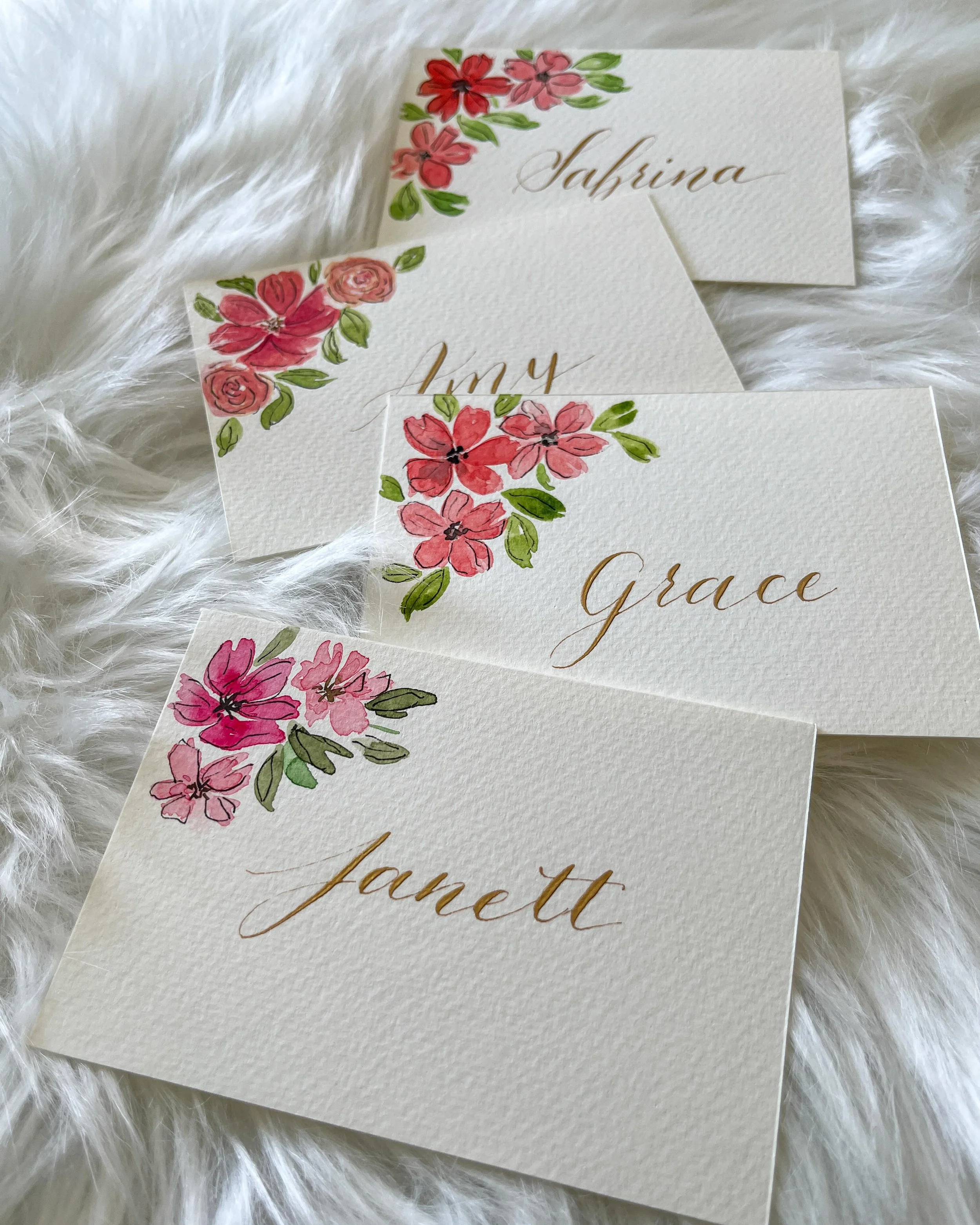 Four white cards with floral watercolor accents and gold cursive names reading Jannet, Grace, Amy, and Sabrina on a white fluffy surface.