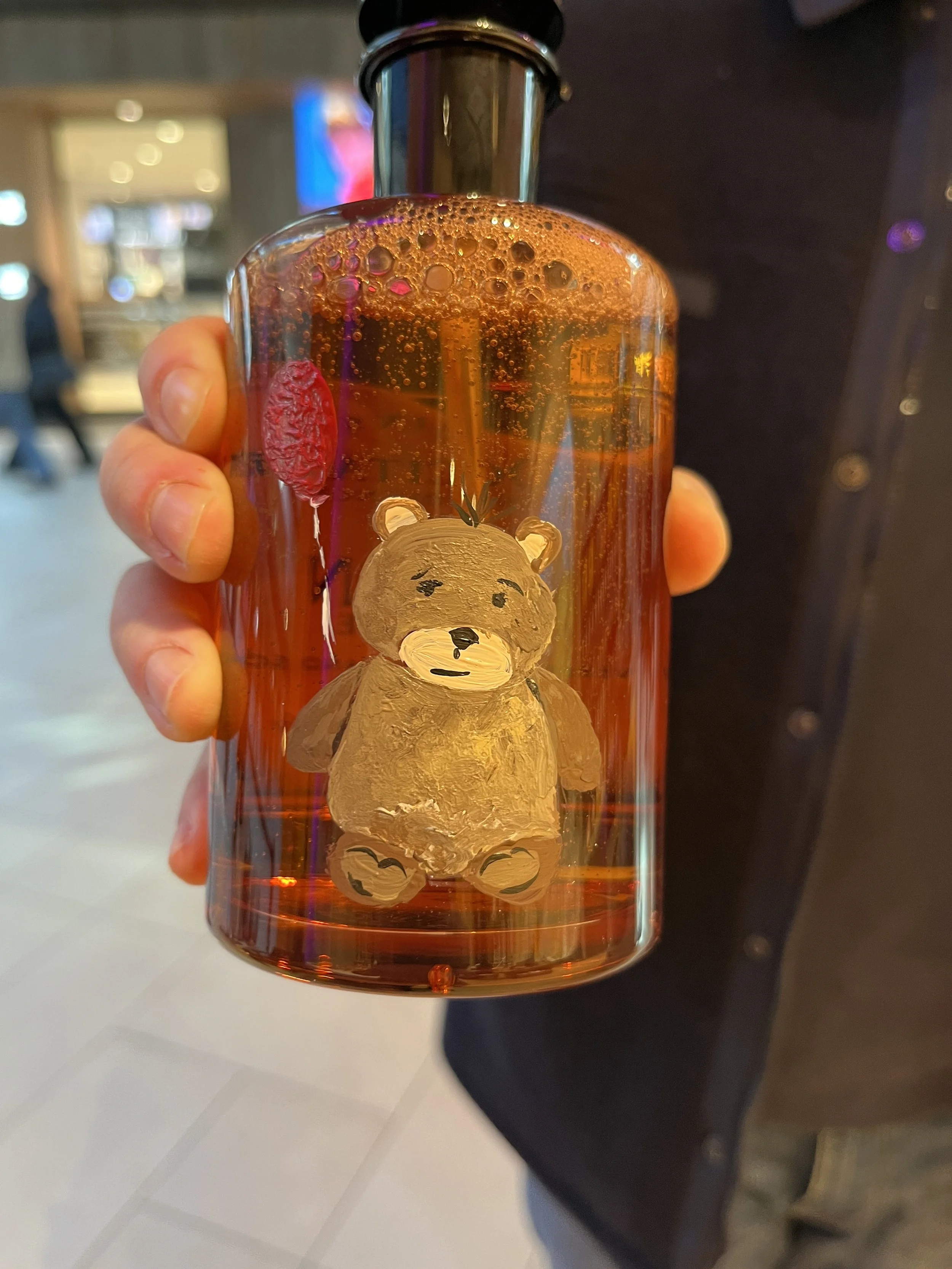 A clear glass bottle with a bear illustration on it, filled with a reddish-brown liquid and foam bubbles on top. The bottle is held by a person with a blurred background featuring a brightly lit indoor setting.