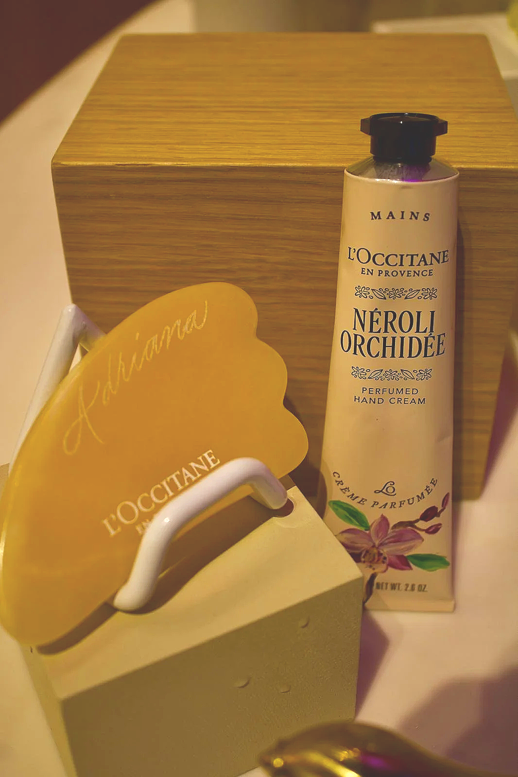 A tube of L'Occitane Neroli Orchidee perfumed hand cream placed next to a gold-colored box and a small container with a white handle, all on a surface.
