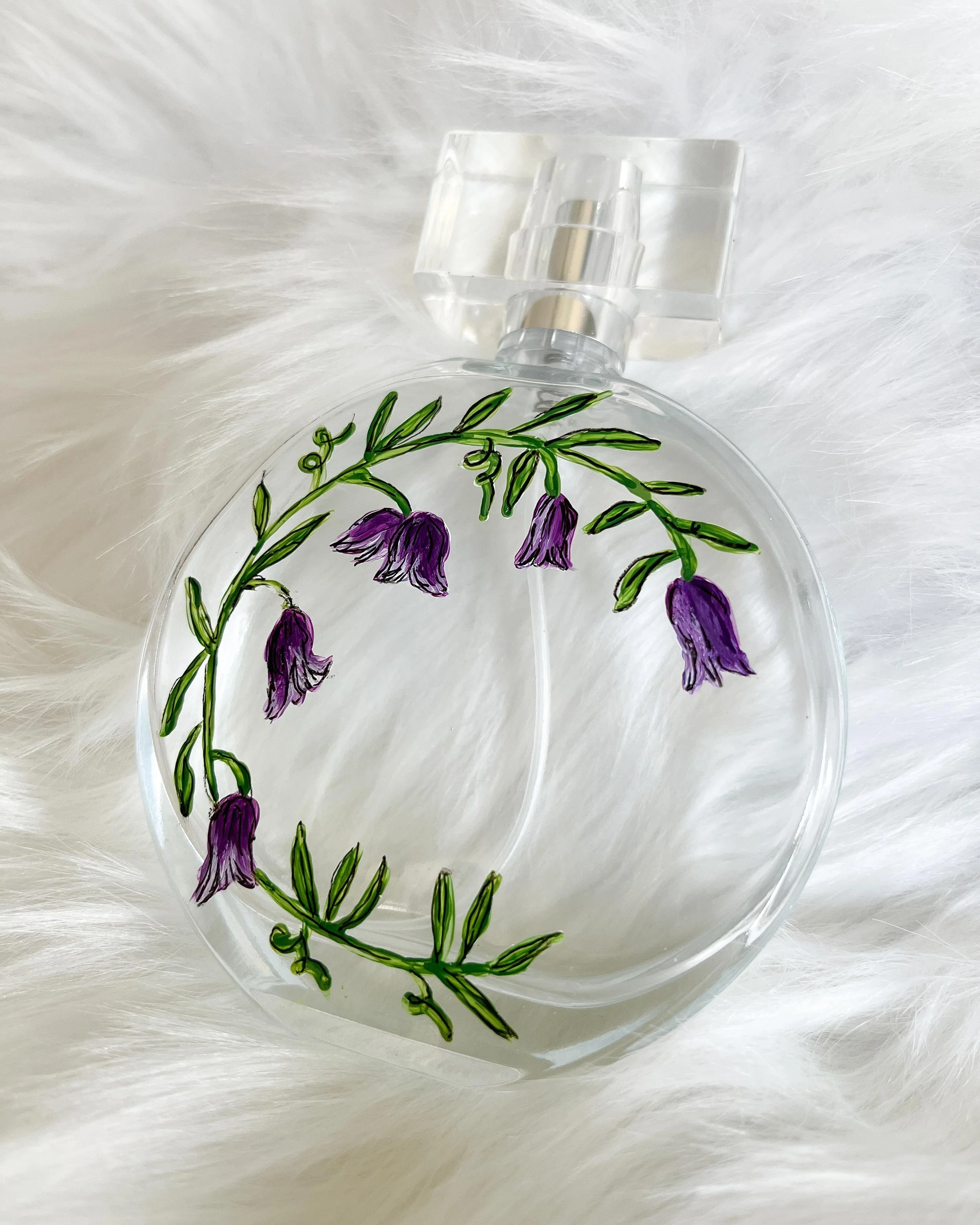 Clear round perfume bottle with a painted purple flower and green leaves design, placed on a white furry surface.
