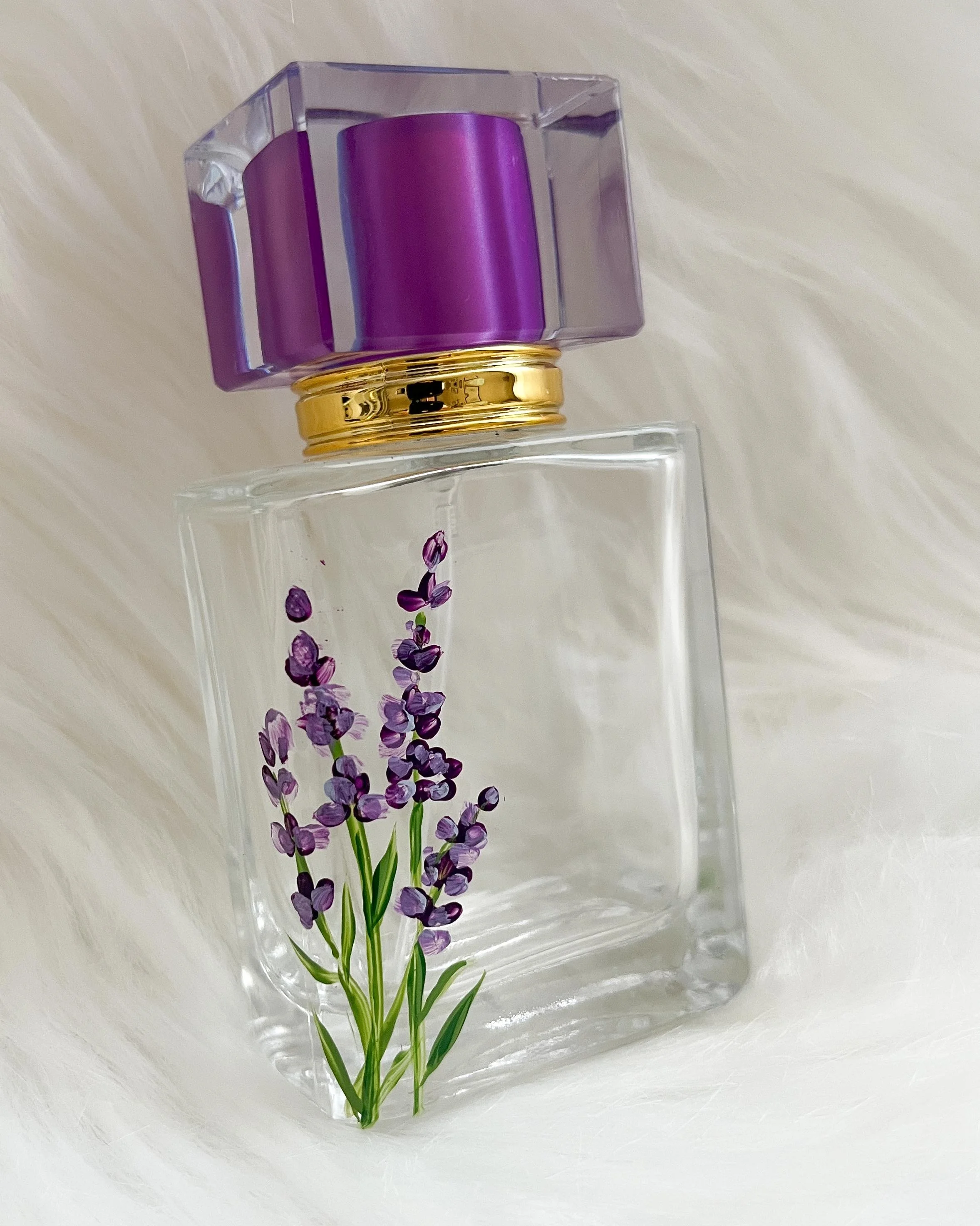 A clear glass perfume bottle with a purple flower design and a purple cap, placed on a white fur surface.